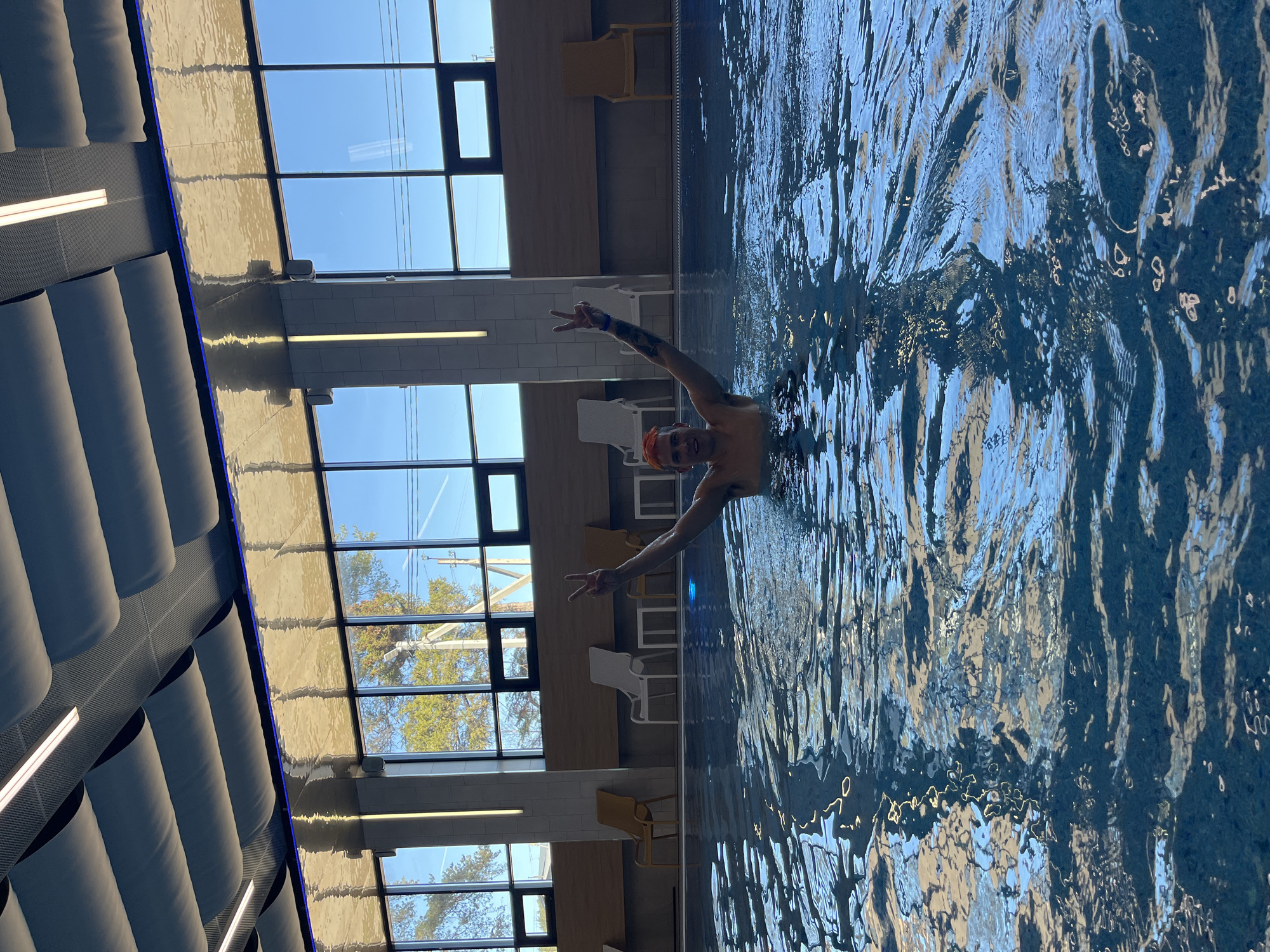A man with short orange hair, wearing a swim cap, is in a swimming pool and giving the peace sign with both hands. He is facing the camera. The indoor pool has large windows with visible trees and power lines outside. There are empty chairs along the side of the pool. The location is an indoor pool with a modern design. There is no way to verify the location in Votkinsk, Russia from this image.