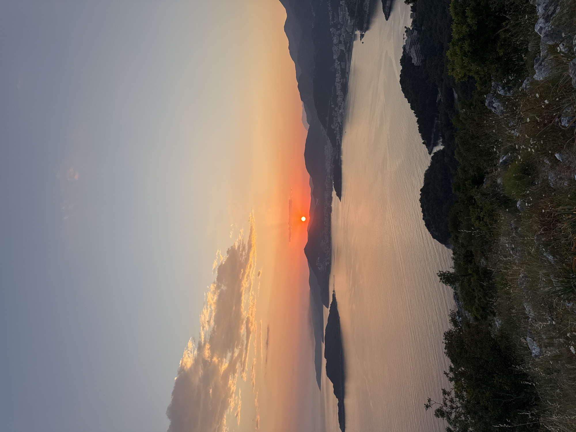 The image captures a sunset over water, likely a lake or sea, with mountainous landmasses on either side. The sun, a bright orange sphere, is positioned near the horizon, partially obscured by a mountain range. The sky transitions from a pale blue in the upper left to shades of orange and yellow closer to the sun. There are some clouds scattered in the lower left portion of the frame, reflecting the sunset colors.
The water is relatively calm, with subtle ripples on the surface, and it reflects the warm tones of the sunset. Landmasses on the right side of the image are covered in greenery, while those on the left appear rocky and barren. The landscape is partially lit, with the areas facing the sun exhibiting brighter highlights, while other areas are in shadow. The location is potentially Sveti Stefan, Montenegro, inferred from the coastal landscape and mountainous backdrop. However, without additional context, this cannot be confirmed with certainty.