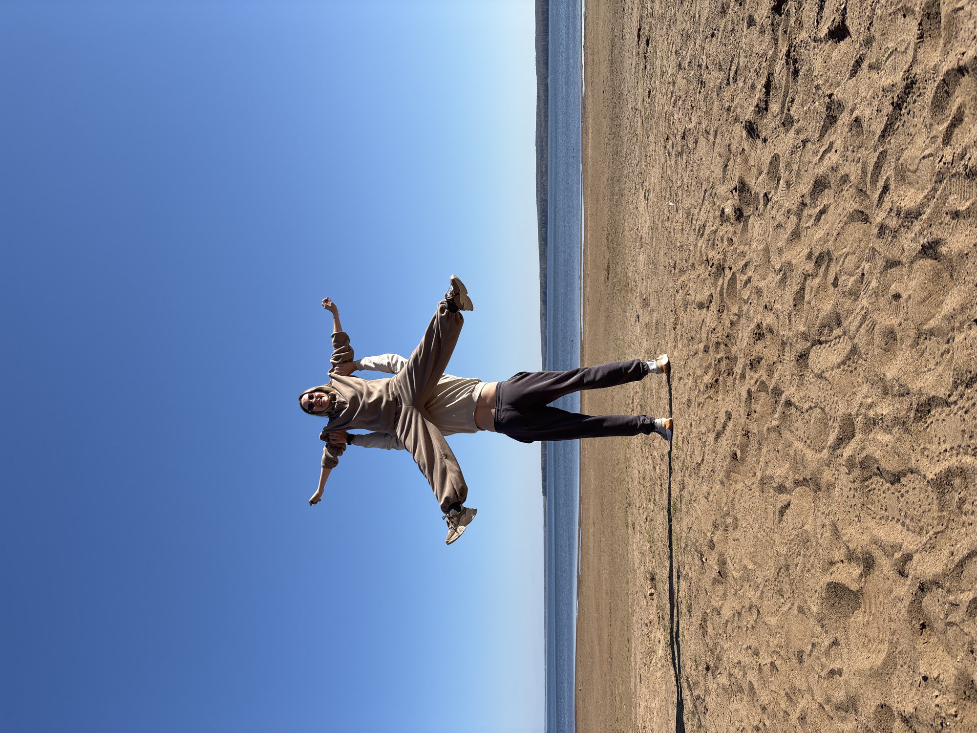 Two individuals are positioned horizontally against a backdrop of a blue sky, a body of water, and a sandy beach. The person on the left is wearing a light brown hoodie and pants, and their arms are outstretched. The individual on the right is dressed in a dark pair of pants. The location appears to be a beach in Chaikovsky, Russia.