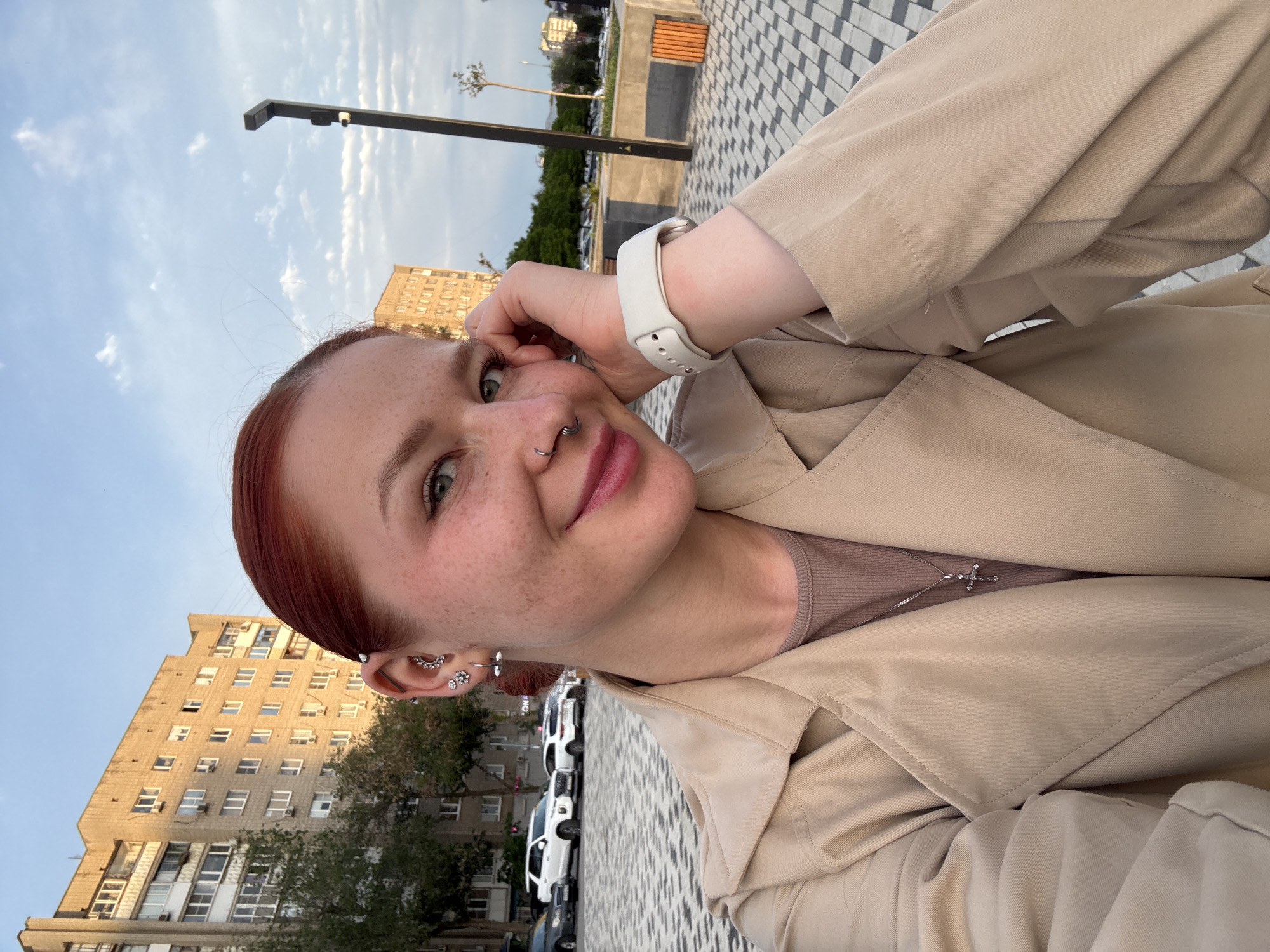 The image is a selfie of a woman with fair skin, freckles, and reddish hair, likely taken outdoors in Rostov-on-Don, Russia, based on the building and street details. She is wearing a light beige trench coat, a light brown top, and a silver cross necklace. Her ear is adorned with multiple piercings, and she has a nose ring. She is wearing a white smartwatch on her left wrist. The woman has a slight smile and her hand is near her face. The background shows a paved area with a patterned design, possibly a sidewalk or promenade. In the distance, there is a street with parked cars, trees, and a tall building with multiple windows, typical of Soviet-era architecture in Russia. A street lamp is also visible. The sky is partially cloudy.
