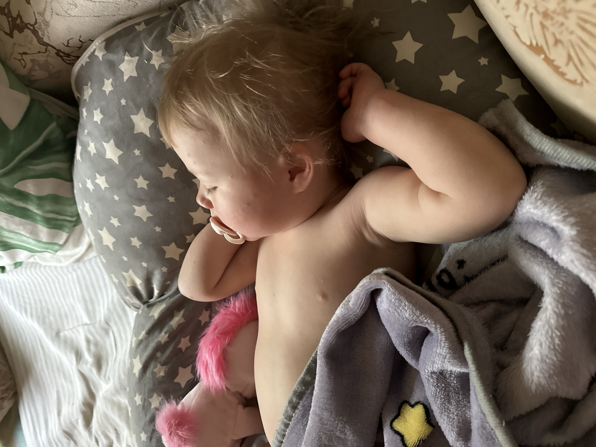 A top-down, medium shot depicts a fair-skinned toddler sleeping on a gray pillow patterned with white stars. The child has light brown, tousled hair. A pacifier is in their mouth. The child is covered partially by a light purple blanket with a yellow star detail. A pink stuffed animal is tucked near their side. The location is unidentifiable.