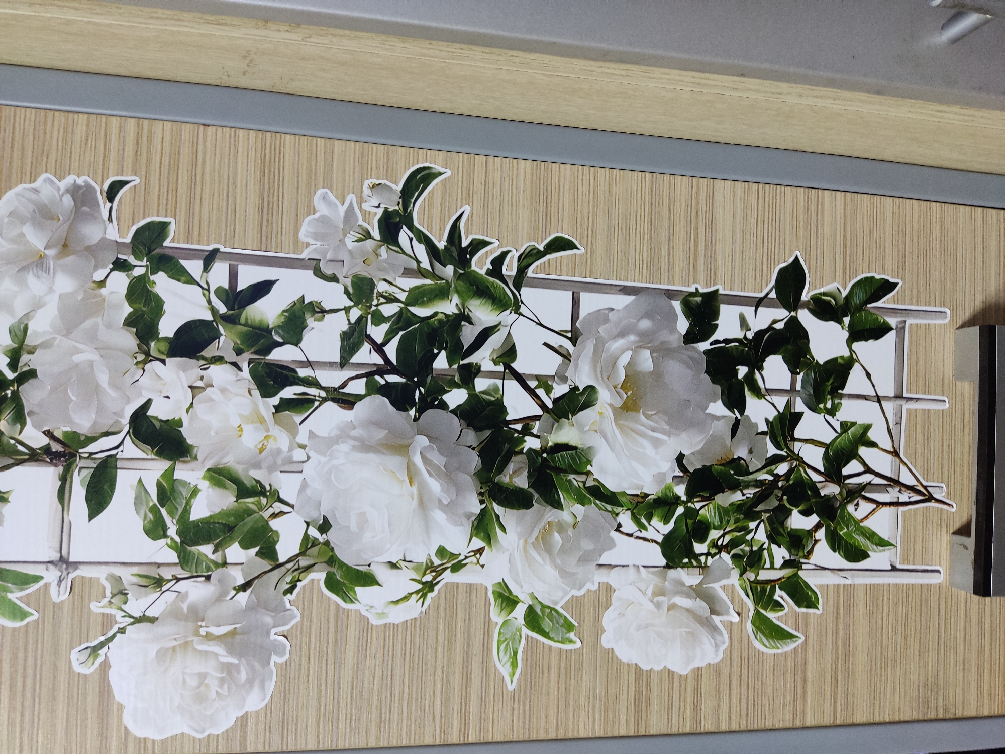 A vertical sticker of white roses and green leaves climbing a white trellis is adhered to a wooden surface. The sticker is positioned against a background of light brown, horizontal wood grain. To the right of the sticker, there is a gray vertical strip, followed by a lighter wood-grain vertical strip. The image does not contain any people or show any specific location within Noginsk, Russia.