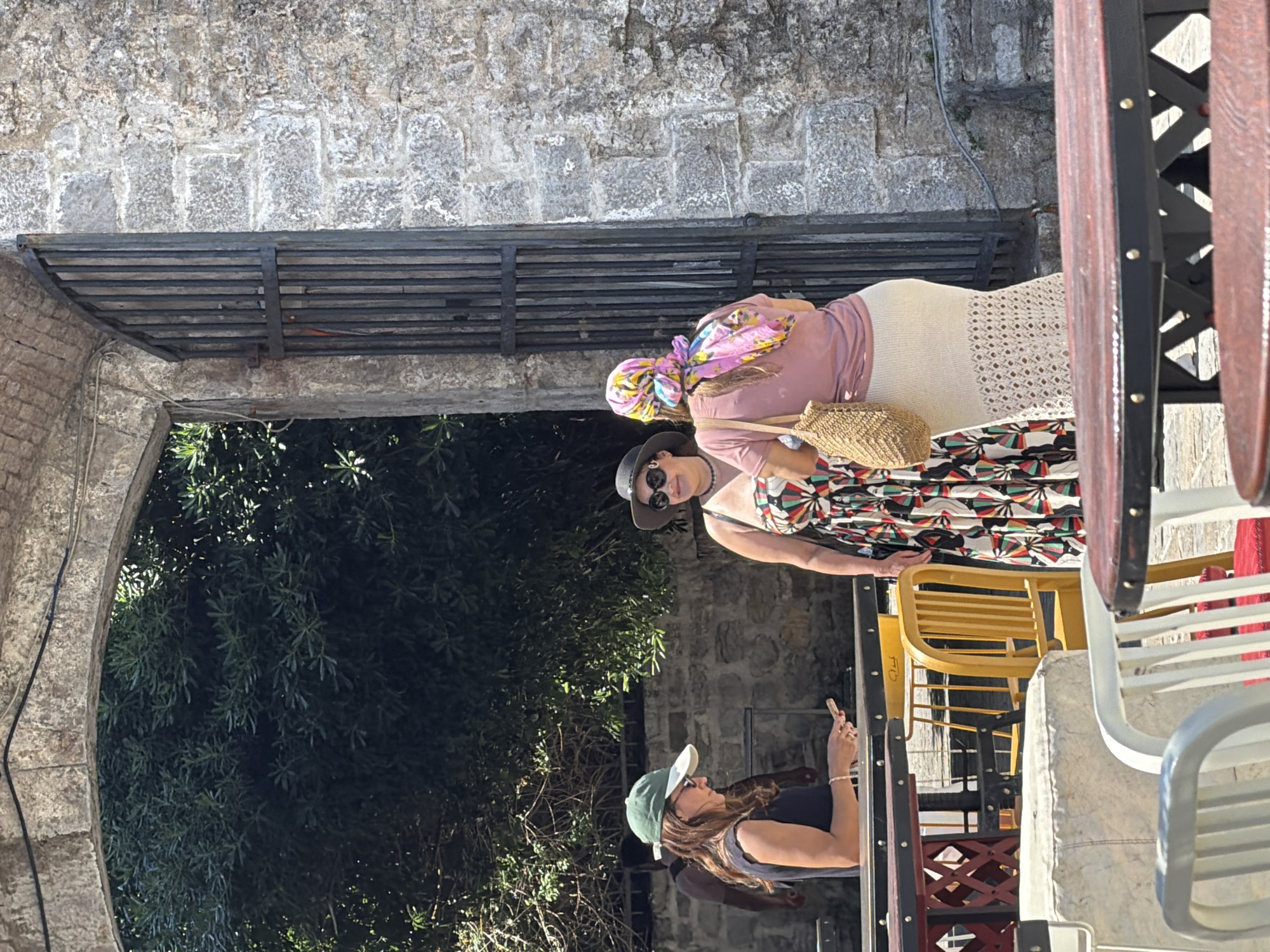 This outdoor image, taken in Budva, Montenegro, depicts three women in what appears to be a cafe or restaurant setting during daytime. The scene is bright and sunny, casting distinct shadows.
In the background, behind the women, there are weathered stone walls and a dark metal grate or shelf structure. Lush green foliage from trees or bushes is visible through an archway on the left side of the frame.
The subjects are three women, casually dressed for warm weather.
* The woman furthest back wears a pink long-sleeved top, a colorful patterned headscarf, and a long, light-colored skirt with a crochet-like pattern. She carries a woven straw bag.
* Standing slightly in front of her is another woman wearing a vibrant, multi-colored patterned dress, a dark hat, and sunglasses.
* In the foreground, to the right, a third woman is visible from the waist up, facing to the right (her left). She wears a dark top, a green baseball cap, and sunglasses, and is holding a smartphone in her right hand, seemingly looking at the screen.
The area is furnished with various outdoor seating options. Several chairs are visible: a bright yellow plastic chair, white metal chairs, and chairs with red accents, along with parts of tables. The overall atmosphere suggests a relaxed, social gathering in a historic or scenic location.
There is no clearly discernible text visible within the image.