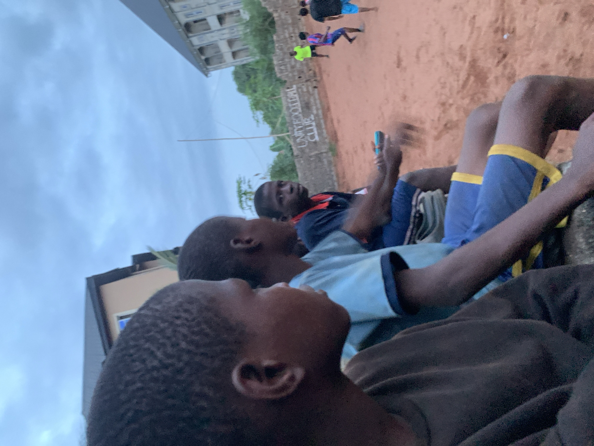 The image shows several boys sitting outside in Ukwu-Abwa, Nigeria, possibly near a soccer field ("UNITED FOOTBALL CLUB" is visible on a wall in the background). Some people are playing soccer on the field in the background. One of the boys is holding a blue device, possibly a phone. The environment is overcast. Buildings and greenery are also visible.