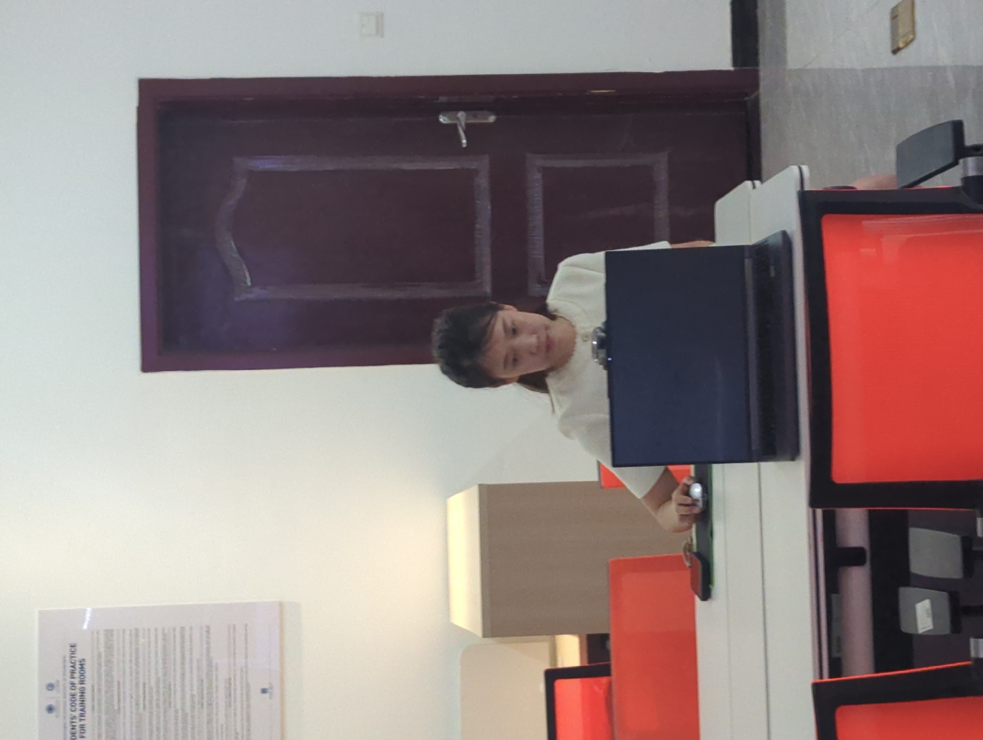 A person sits at a desk with a computer. The room has a door, a document posted on the wall, and orange chairs. The location is Jiangjin, China.
