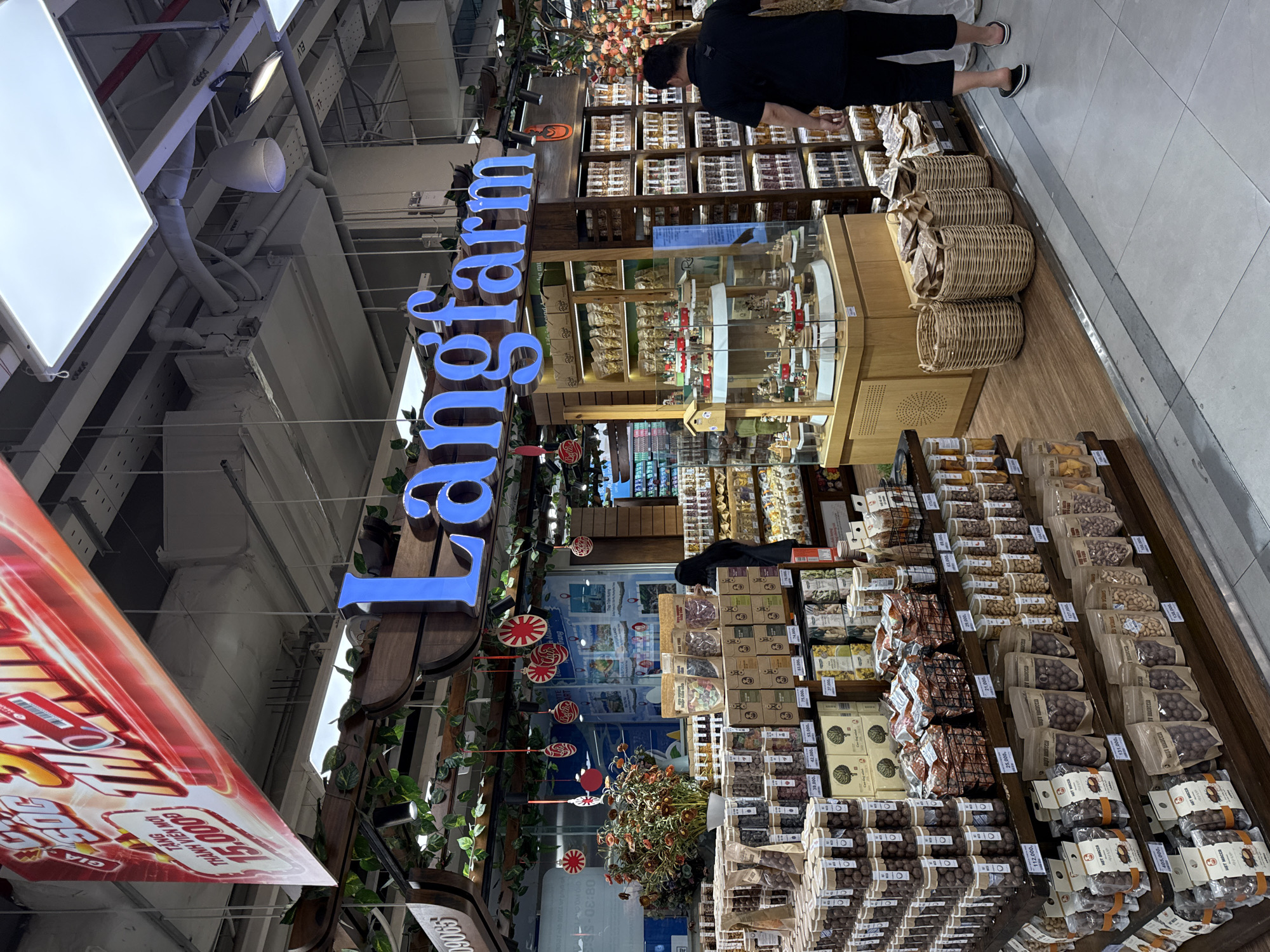 The image shows an interior view of a retail store, likely a supermarket or a specialty food shop, with a prominent "Langfarm" sign in blue illuminated lettering. The store appears well-stocked with various food products, primarily packaged nuts, dried fruits, and possibly candies, displayed on wooden shelves and in baskets. There are also decorative elements like artificial plants and hanging ornaments.
Two individuals are visible in the scene. One person, wearing a black shirt and black pants, is seen from the back, facing towards the right side of the image and appearing to walk away. Another person, wearing a black top and a dark skirt, is partially visible in the middle of the frame, facing towards the left.
The store's lighting is bright, with overhead fluorescent lights and spotlights illuminating the merchandise. The flooring is tiled, and a low metal barrier is visible in the foreground. A large red banner with Vietnamese text and graphics is hanging from the ceiling in the upper left portion of the image.
Based on the "Langfarm" branding, which is a well-known Vietnamese brand specializing in agricultural products and processed foods, and the general appearance of the store, it is plausible that this location is within a modern shopping center or supermarket in Nha Trang, Vietnam. The products displayed, such as dried fruits and nuts, are consistent with the offerings of Langfarm. The overall atmosphere suggests a commercial retail environment focused on food sales.
