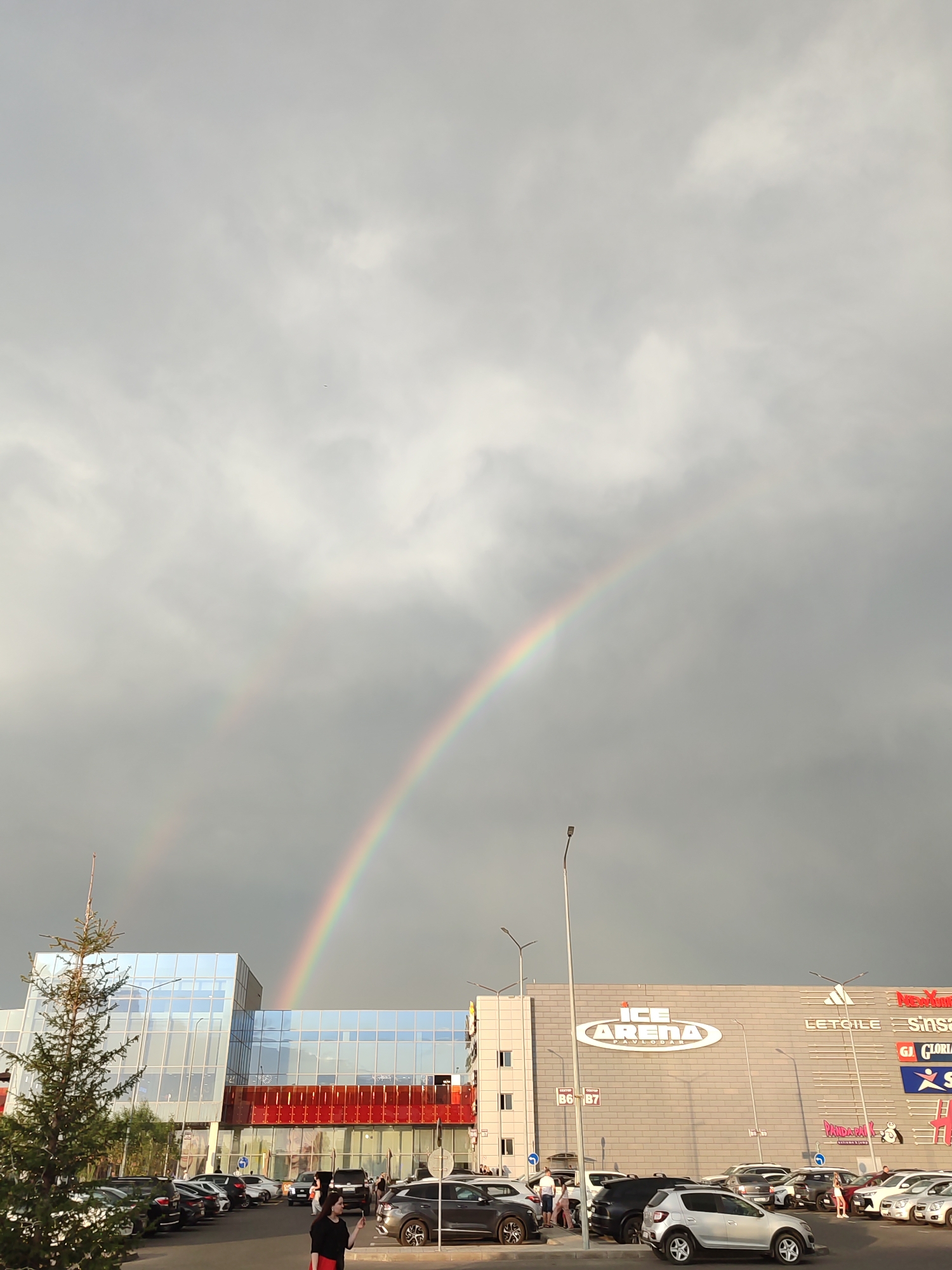 The image captures a cloudy sky over a commercial building in Pavlodar, Kazakhstan, with a visible double rainbow. The building, identified as "ICE ARENA PAVLO DAR," features a modern facade with large glass windows and red accents. Several cars are parked in a lot in front of the building, and a few people are visible near the vehicles and on the sidewalk. A coniferous tree stands to the left of the building. The overall scene suggests an outdoor environment, possibly a shopping center or entertainment complex, under a sky indicative of recent or impending rain.