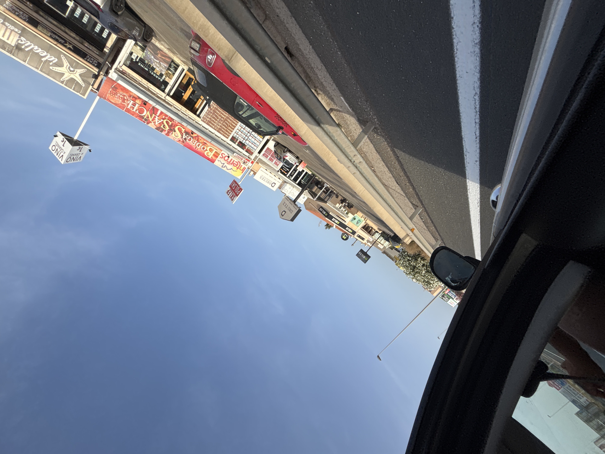 The image shows a road scene in Urbanización Cabo Roig, Spain, with a bright blue sky overhead. The perspective is from inside a vehicle, looking outwards. A metal guardrail runs along the side of the road. Beyond the guardrail, there are several shops and businesses with visible signage. One sign reads "Bodegas SANCH", another has "VINO A CHANCE 1€" on a cube-shaped structure. There are red and grey cars parked or driving along the road. The buildings appear to be low-rise commercial structures. In the foreground, the interior of the vehicle is visible, including a side mirror and part of the dashboard. The lighting suggests it is daytime, likely late afternoon due to the angle of the shadows.