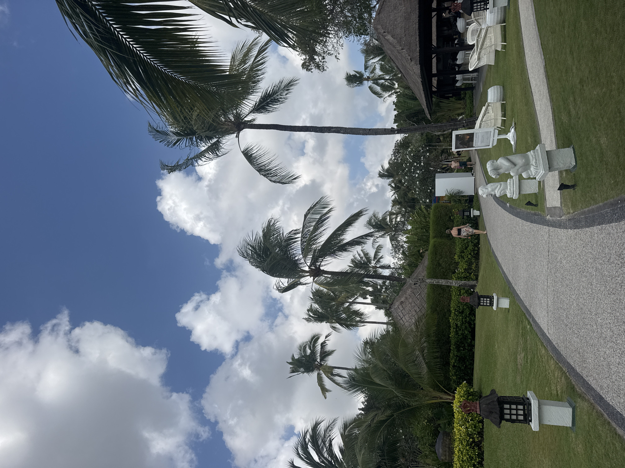 This is a medium-shot, eye-level, vertical, outdoor photograph taken in the city of Kuta Selatan, Indonesia. The scene depicts a resort or hotel pathway with lush green grass on either side, lined with palm trees and tropical vegetation. The sky is a vibrant blue with scattered white clouds.
In the foreground, a light-colored, pebble-textured pathway curves from the bottom right towards the upper left. On the left side of the path, two dark-colored, square lanterns with thatched roofs sit atop white pedestals. Further down the path, a woman wearing a black top and a patterned skirt walks away from the camera. Two white, stone sculptures of seated monkeys on pedestals are visible on the grass to the right of the path.
In the mid-ground, the pathway continues to curve. A tall, slender palm tree stands prominently on the right side. Beyond the path, there are thatched-roof structures, including what appears to be a bar or restaurant area with white wicker seating arranged on the grass. Several other palm trees and tropical plants fill the background, with dense green hedges bordering a portion of the landscape. The overall impression is one of a serene and tropical resort environment.