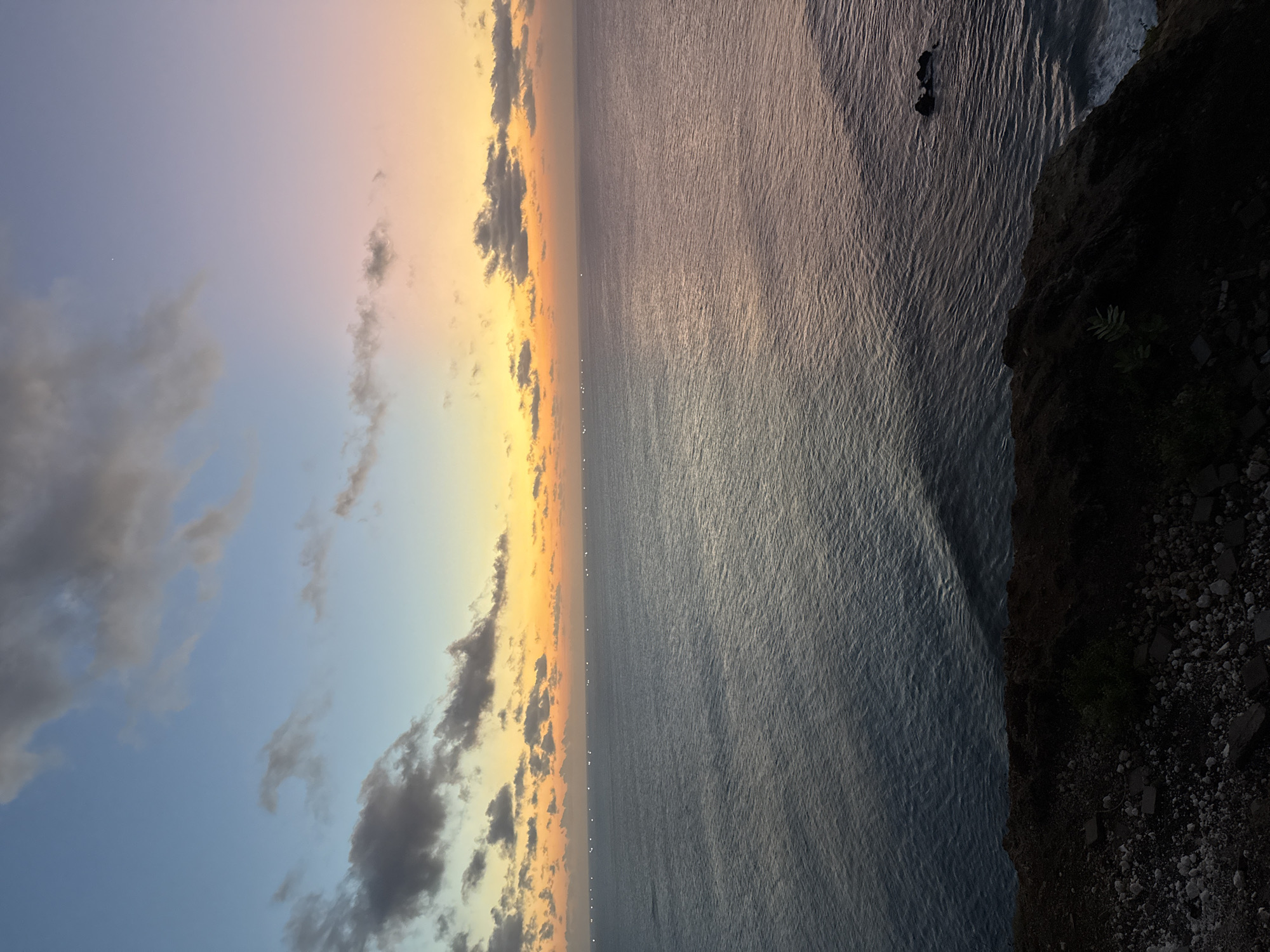 An ocean view from a cliff overlooking the water at dawn. The sky displays a gradient of colors from light blue at the top to yellow, orange, and pink near the horizon. Clouds are scattered across the sky. The ocean is calm with gentle ripples. A line of small, distant lights is visible on the water's surface, stretching across the horizon. A dark, rocky cliff edge occupies the foreground, with some sparse vegetation. A few dark rocks are visible in the water to the right. The scene captures the peaceful transition from night to day over the ocean. The location is Kuta Selatan, Indonesia.