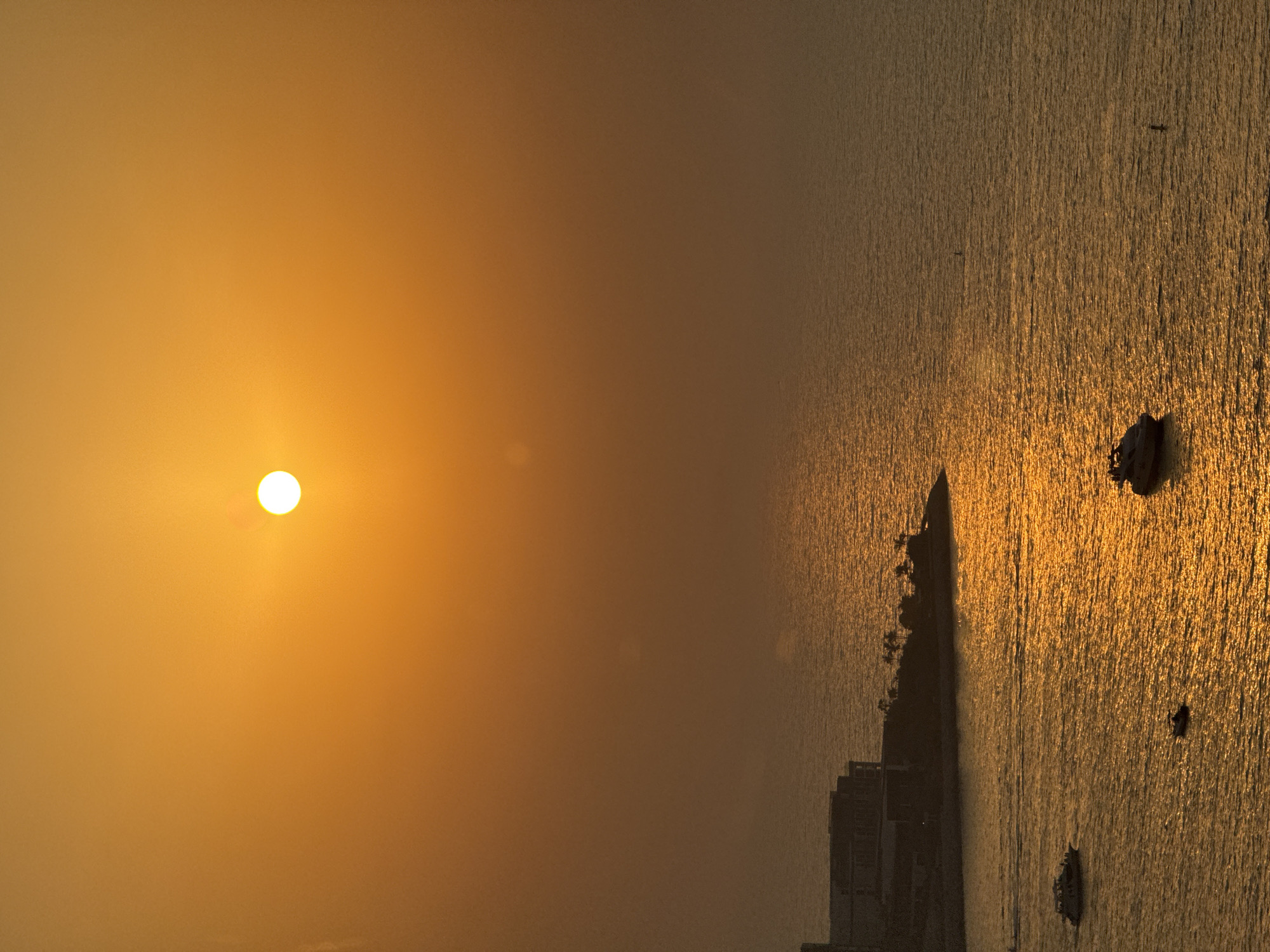 This is an image of a sunset over the Arabian Gulf in Dubai, United Arab Emirates. The sun, a bright white disk, is positioned in the upper center of the frame, casting a warm, orange glow across the sky and the water. In the foreground, a collection of boats are visible on the shimmering, golden surface of the sea. To the left, a modern building complex, likely a hotel or residential area, sits on a man-made peninsula jutting out into the water. The architecture appears sleek and contemporary, with multiple levels and dark facades silhouetted against the vibrant sunset. The water reflects the intense colors of the sky, creating a dazzling effect. The overall scene evokes a sense of tranquility and luxury, characteristic of Dubai's coastal landscape.