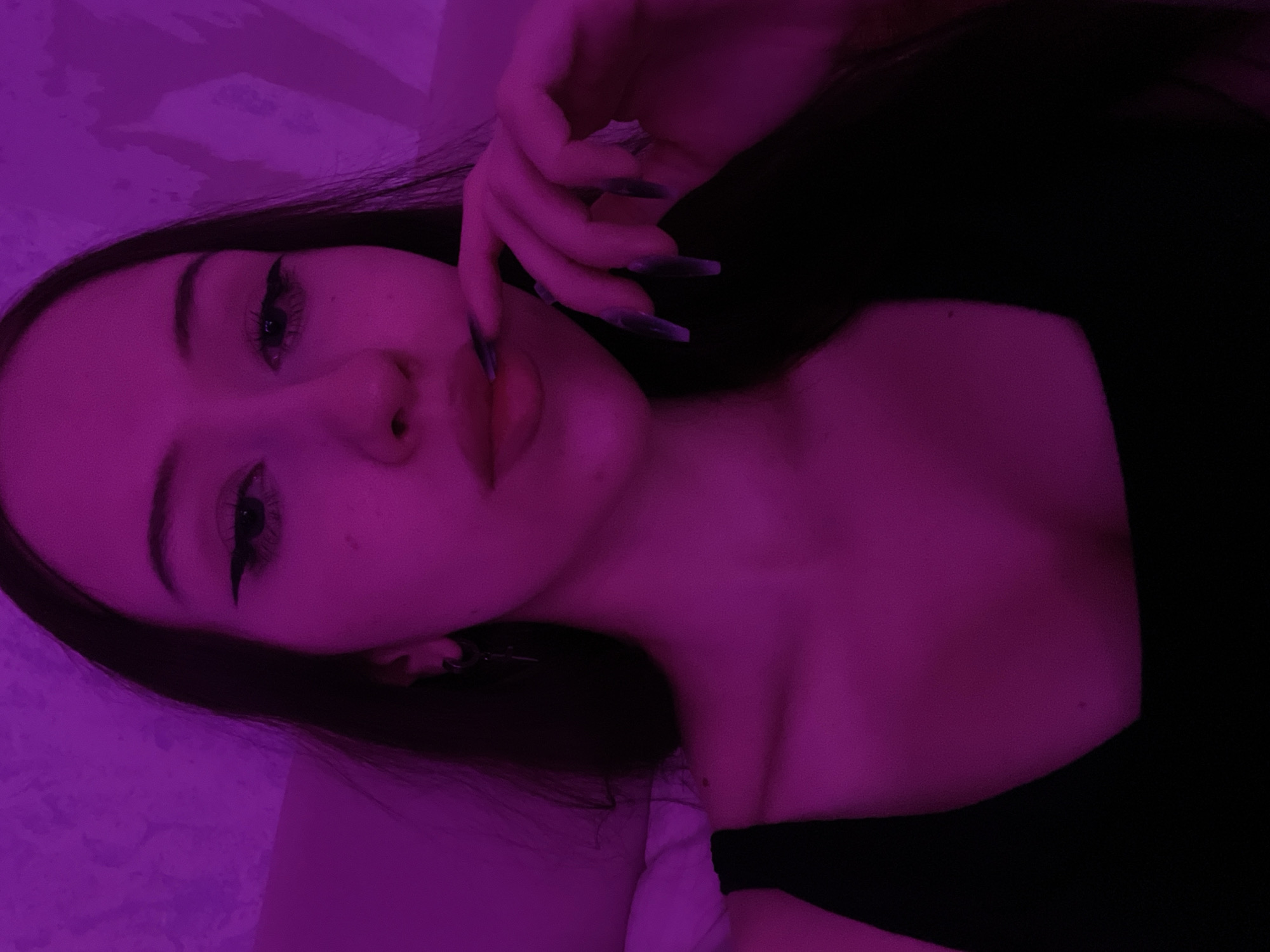 The image is a selfie taken in a dimly lit room with purple lighting. The subject is a young woman with long, dark hair and noticeable winged eyeliner. She is wearing a black top. Her right hand is brought to her mouth, with long, pointed fingernails visible. The background appears to be a textured wall. No specific location in Votkinsk, Russia can be determined from the image.
