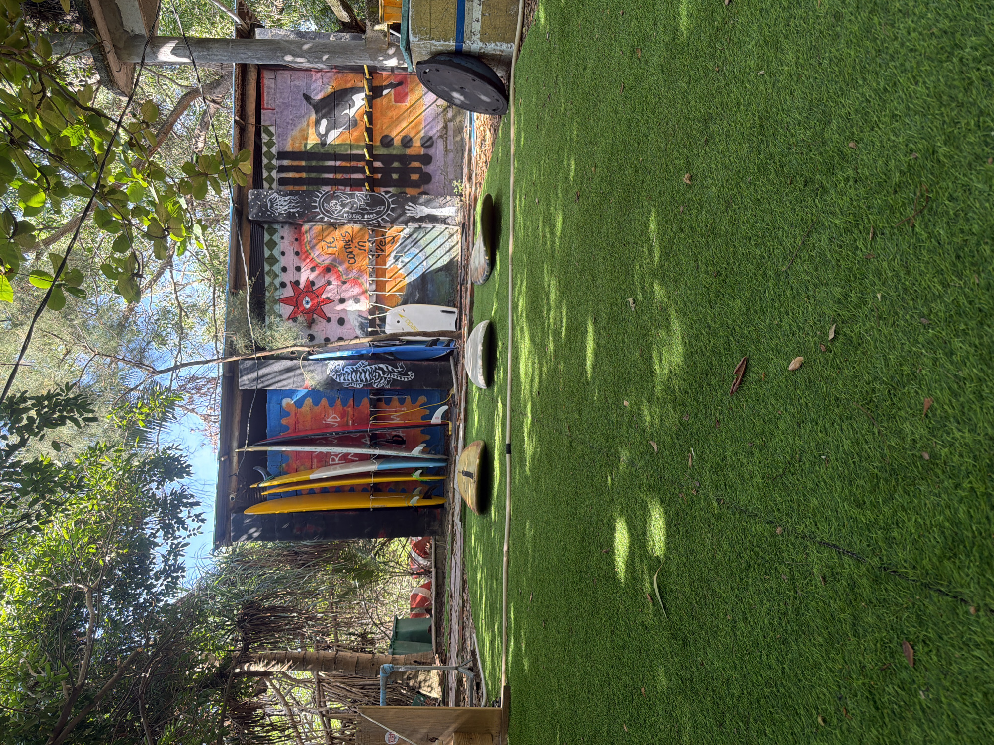 The image shows an outdoor scene in Cam Lam, Vietnam, featuring a wooden shed with surfboards leaned against it. The shed is adorned with various graffiti and murals. In front of the shed is an area covered with artificial turf. A few round, stepping-stone-like objects are placed on the turf. The shed is partially obscured by trees and foliage.