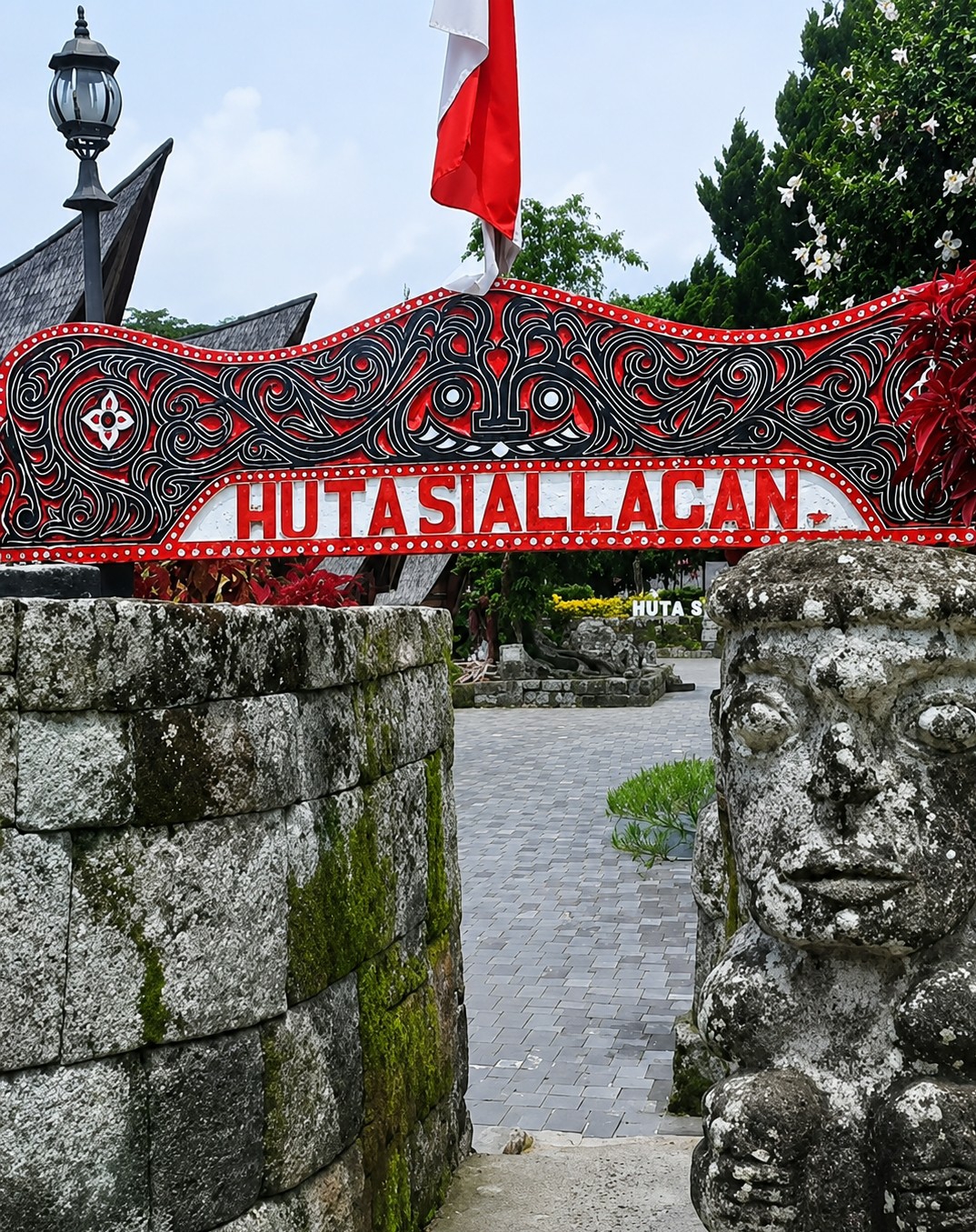 The image depicts the entrance to Hutasiallagan in Ambarita, Indonesia. A decorative red and black archway with white lettering spelling "HUTASIALLAGAN" spans the foreground. To the left, a stone wall covered in moss stands beside a lamppost and a portion of a traditional roof. To the right, a weathered stone statue with a human-like face is partially visible, also showing signs of moss growth. Beyond the archway, a paved courtyard is visible, leading further into the site, with trees and foliage in the background. A red and white flag is prominently displayed above the archway. The sky is overcast with clouds.
