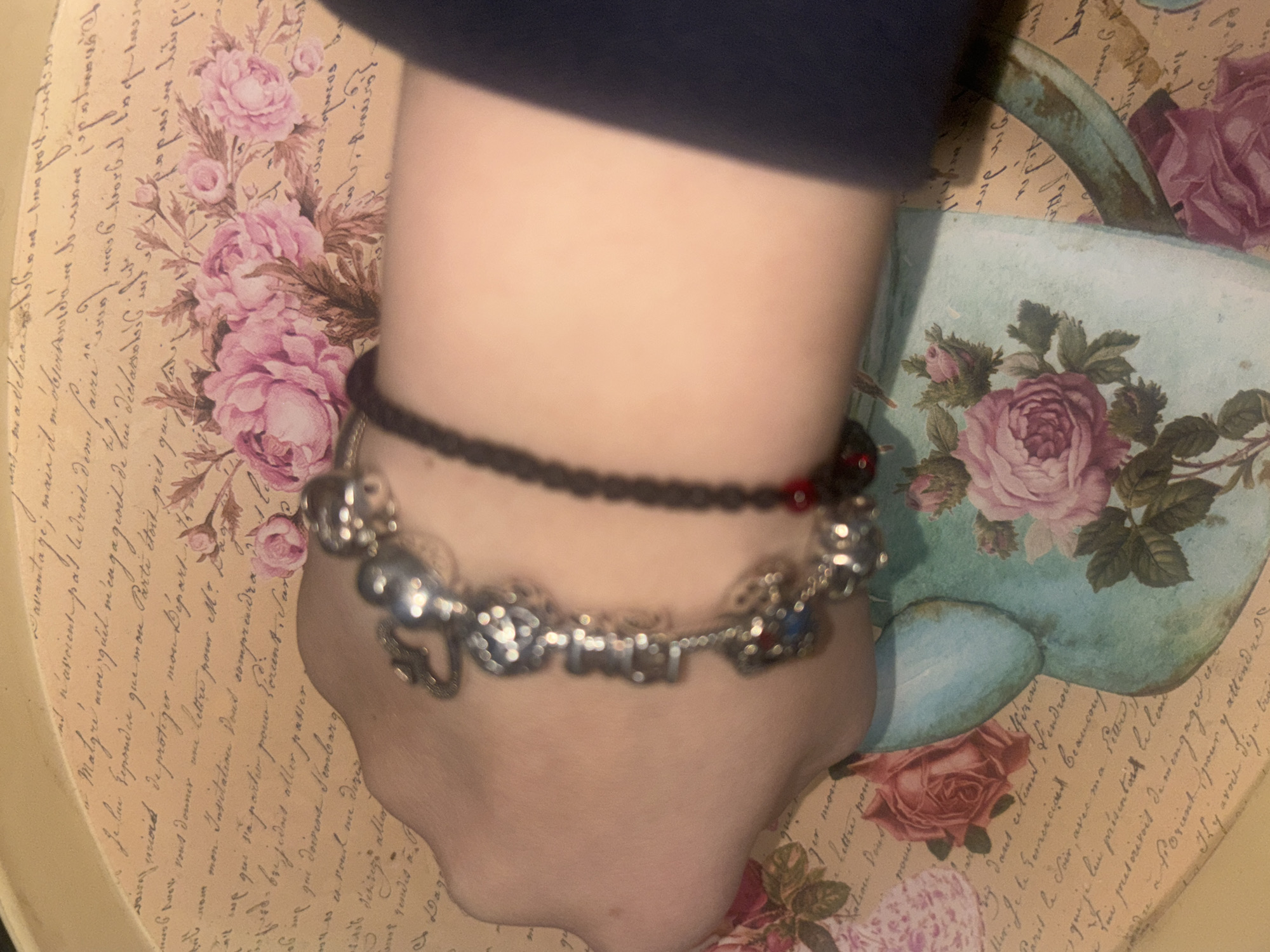 The image shows a wrist with two bracelets against a background of a decorative tray with floral and script designs. The wrist appears to be that of a person wearing a dark-sleeved garment. The bracelets consist of a dark beaded one with occasional red beads and a silver charm bracelet. The tray depicts various pink roses and what looks like a watering can filled with roses against a backdrop of cursive script.
There is no information in the image that places it in Gorni Bogrov, Bulgaria, or any other specific location.