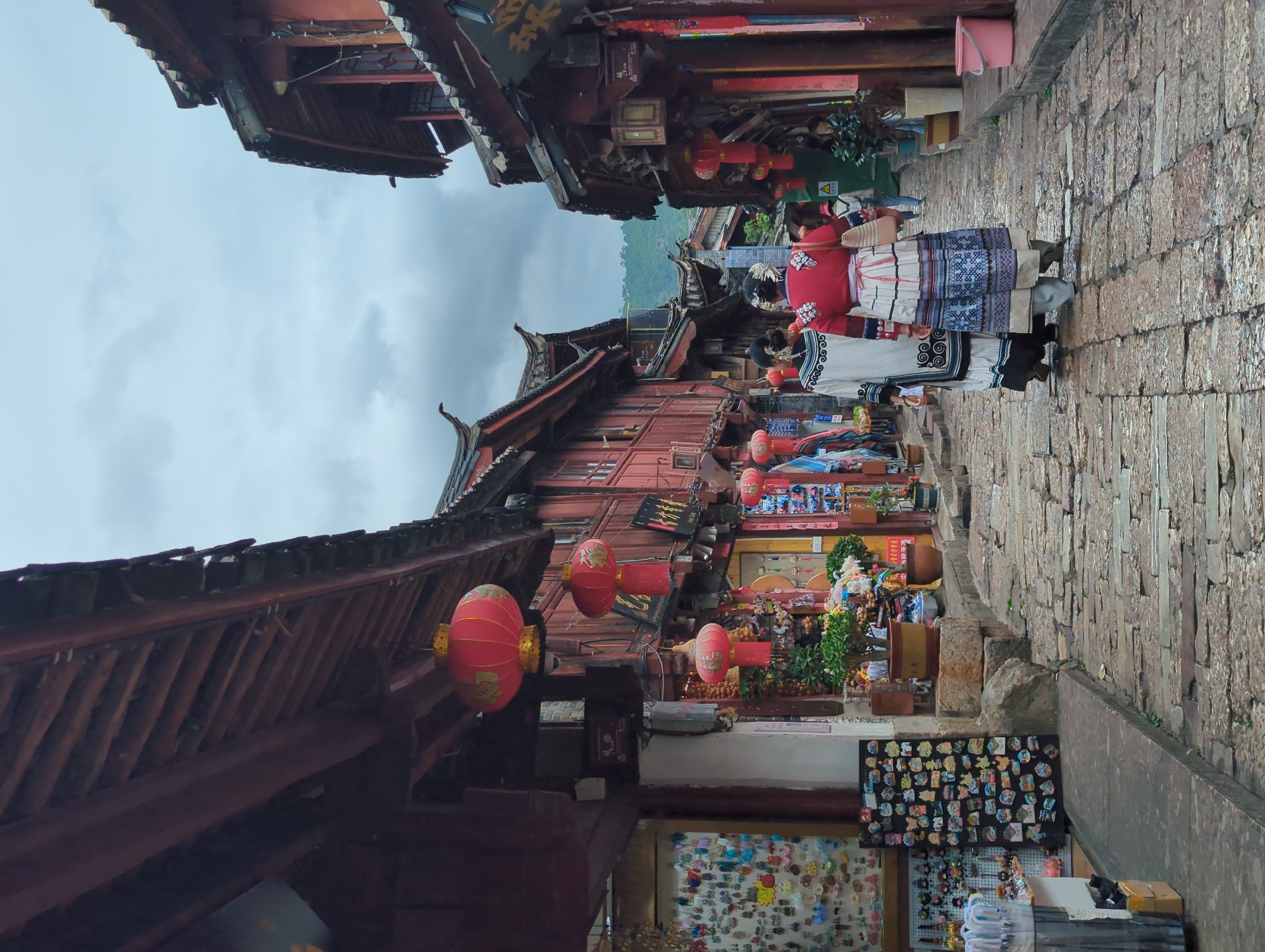 The image depicts a street scene in a traditional Chinese town, likely Lijiang, given the architectural style. The perspective is vertical, as if the camera were held sideways. Red, multi-story buildings with traditional tiled roofs line a narrow, cobblestone street. Red lanterns are suspended from the eaves of the buildings, adding a festive and cultural element.
Several people are visible, dressed in what appears to be traditional ethnic attire. One individual in the foreground wears a red top and a patterned skirt. Another person beside them wears a white top with intricate black and white patterns. They are walking along the street, suggesting movement and daily life.
The street is lined with shops showcasing various goods. Shelves display an array of items, including what look like souvenirs or local crafts such as magnets and figurines. The shops appear to be open, with merchandise displayed outside.
The weather seems overcast, with a cloudy sky dominating the upper portion of the image. The lighting is diffused, suggesting it might be late afternoon or early morning, or simply a cloudy day. The overall atmosphere is one of a bustling historic marketplace, with a strong sense of local culture and tradition. No discernable text is legible in the image.