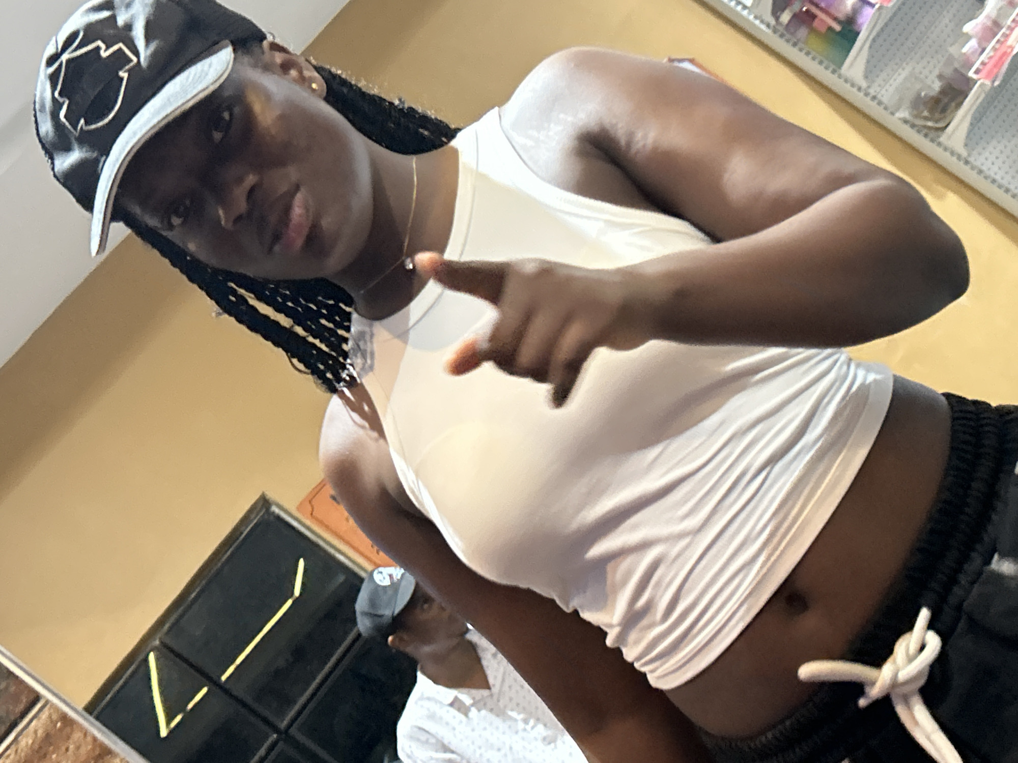 A Black woman wearing a white tank top and black sweatpants with a white drawstring is posing for a photo in a store located in Ihiagwa, Nigeria. She is wearing a black baseball cap with a white logo. Her left arm is bent, and her index finger is pointed towards the camera. Behind her, a Black man wearing a white shirt with a pattern of small grey dots and a grey baseball cap is visible. The background of the image shows a yellow wall and a display shelf with various items.