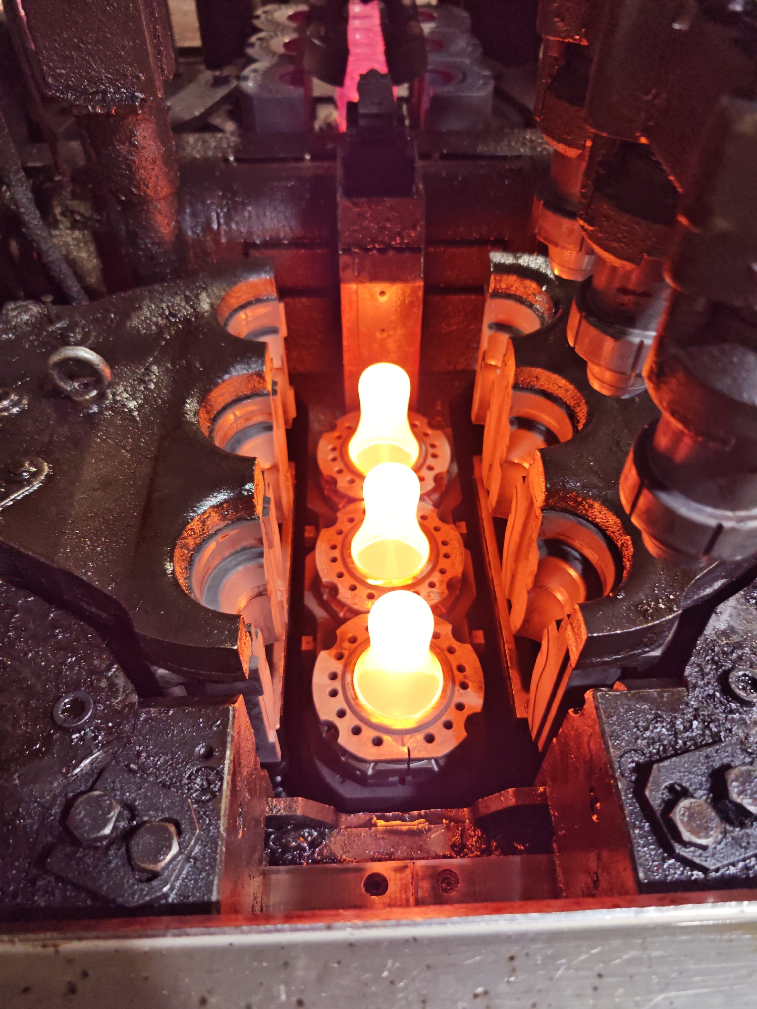 The image depicts a close-up, high-angle view of what appears to be a glass-blowing machine, possibly located in a manufacturing or industrial setting. Three glowing, molten glass shapes are centrally positioned, each held within a ring-shaped fixture. The machinery surrounding the glass is heavily greased and mechanically complex, with visible bolts, gears, and metal components. The lighting is predominantly warm, casting a reddish-orange glow from the molten glass and reflecting off the metal surfaces. The environment appears gritty and industrial. There is no evidence of people present in the image. There is no information available to confirm the location in the city of Pleven, Bulgaria.
