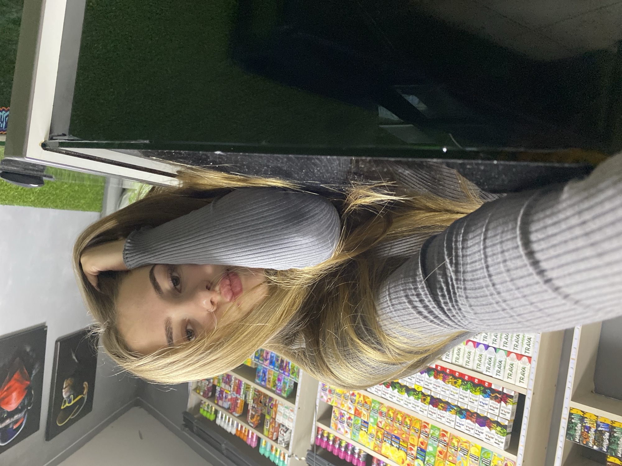 In a store stocked with vape products, a young woman with blonde hair lies on a dark, reflective surface, possibly a counter or table. She is wearing a grey, ribbed long-sleeved shirt. Her head is propped up on her arm, and she is looking towards the camera with her lips slightly pursed. In the background, shelves are filled with colorful vape juice bottles and packaging. To the right of the woman, two framed artworks of monkeys are visible on a grey wall. The overall lighting is bright, illuminating the merchandise and the woman's face. The location is specified as Dyakovo, Russia.