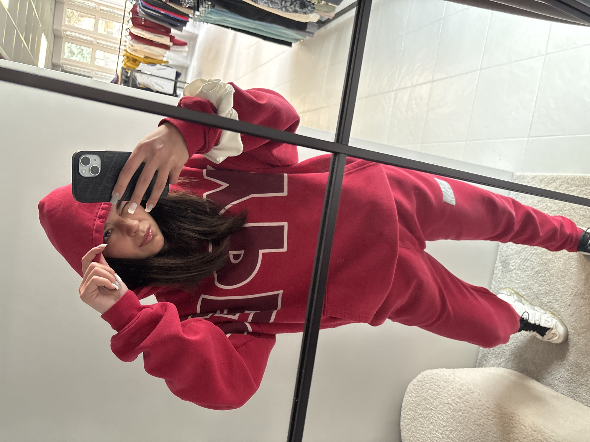 A young woman is taking a selfie in a mirror. She is wearing a red hoodie and sweatpants. The hoodie has white lettering on the front. She is holding a black smartphone in her left hand. The woman has long dark hair and is wearing light nail polish. The reflection in the mirror shows a glimpse of a closet with hanging clothes, a tiled floor, and a white chair or rug. The location is not identifiable as Gabrovo, Bulgaria from this image alone.
