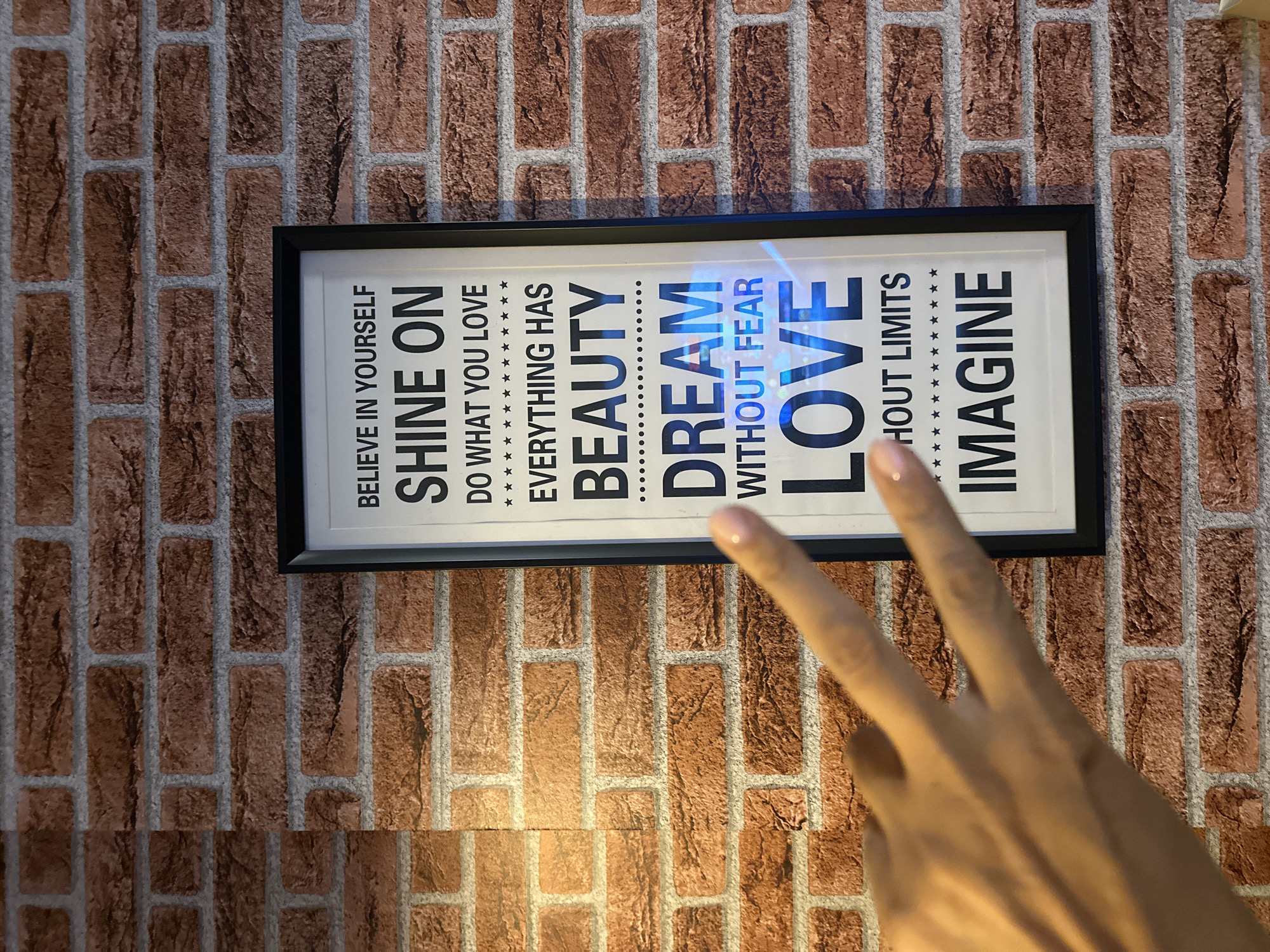 A framed inspirational quote is displayed on a brick-patterned wallpaper. The quote reads: "BELIEVE IN YOURSELF SHINE ON DO WHAT YOU LOVE EVERYTHING HAS BEAUTY DREAM WITHOUT FEAR LOVE WITHOUT LIMITS IMAGINE." A hand with painted fingernails is visible in the foreground, making a peace sign. The scene appears to be indoors, with the brick wallpaper suggesting a decorative element in a room. There is no specific information about the location in Kathu, Thailand.