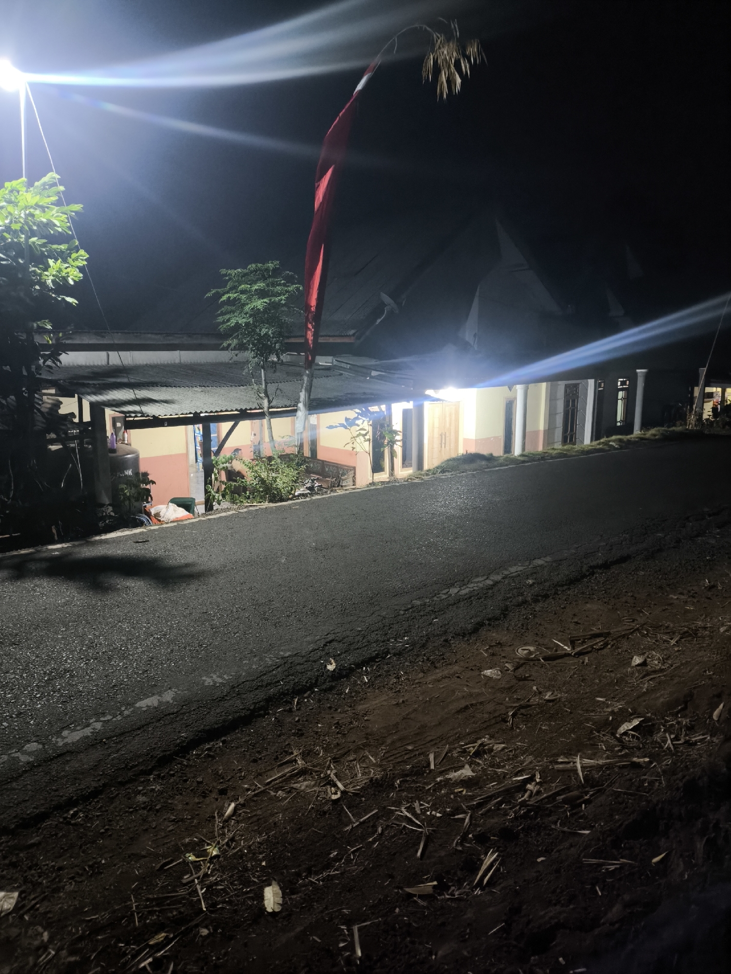 The image shows a dimly lit street scene at night in Bodang, Indonesia. A road, likely paved with asphalt, runs diagonally across the foreground. To the left of the road is a dirt embankment littered with debris, including branches, leaves, and what appear to be discarded plastic items. The right side of the road features a building with a covered porch area supported by columns. The building's walls are painted in light yellow and peach tones, with windows and doorways visible. Trees and foliage are present, with some illuminated by artificial light. A red and white flag, possibly the Indonesian flag, hangs from a pole near the building. Bright artificial lights, likely streetlights, create strong glare and lens flares in the upper left and right portions of the image, obscuring details of the sky and upper structures. The overall impression is of a rural or semi-urban residential area at night.