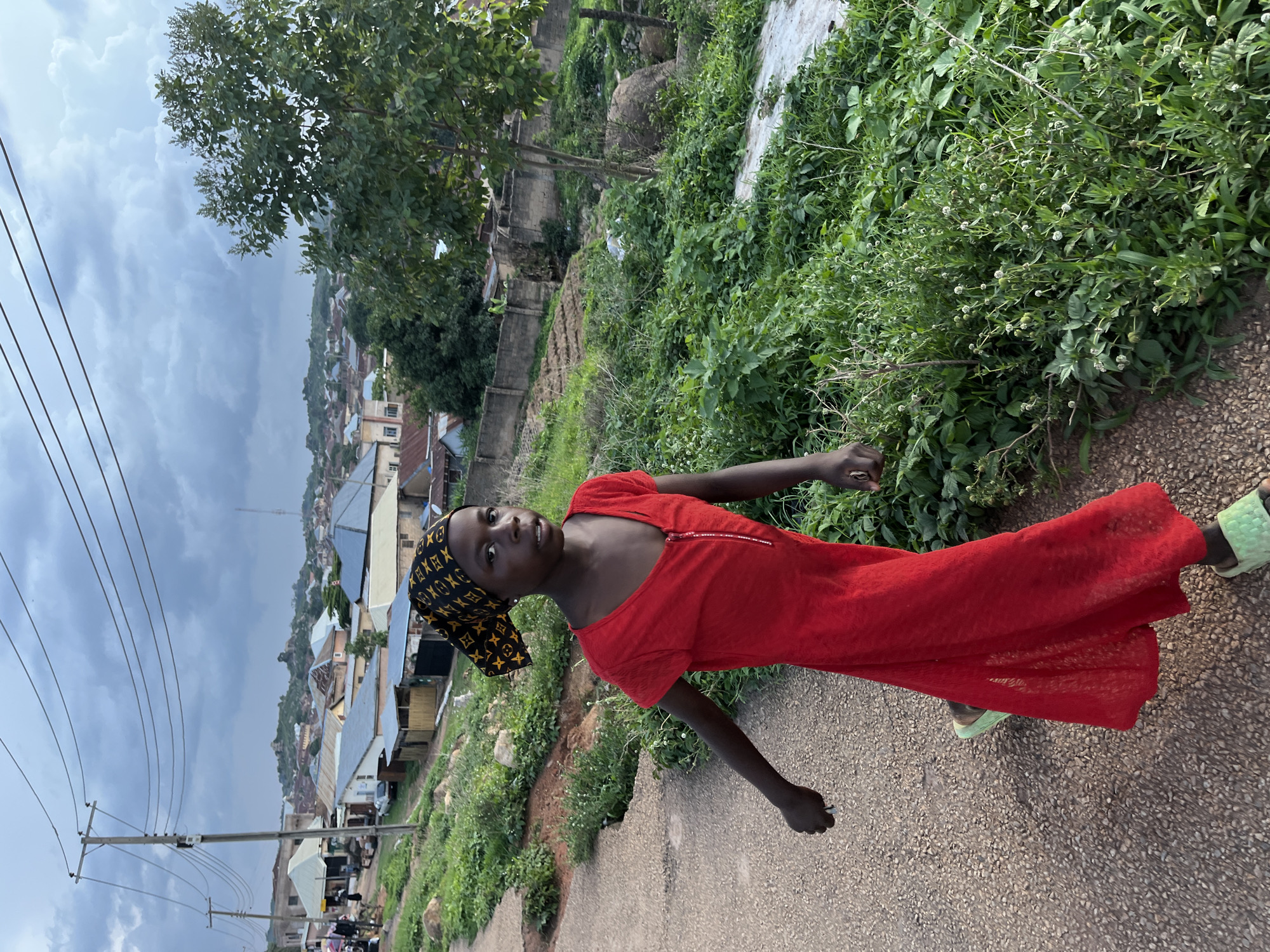 In Jos, Nigeria, a young girl with dark skin walks along a dirt path. She is wearing a red dress with a zipper and a headscarf patterned with gold logos. The girl is walking towards the viewer's left. Behind her, the landscape is hilly and dotted with buildings, some with corrugated metal roofs. Power lines crisscross the sky, and there is lush green vegetation on either side of the path, with some rocks and a large tree visible. The sky is cloudy.