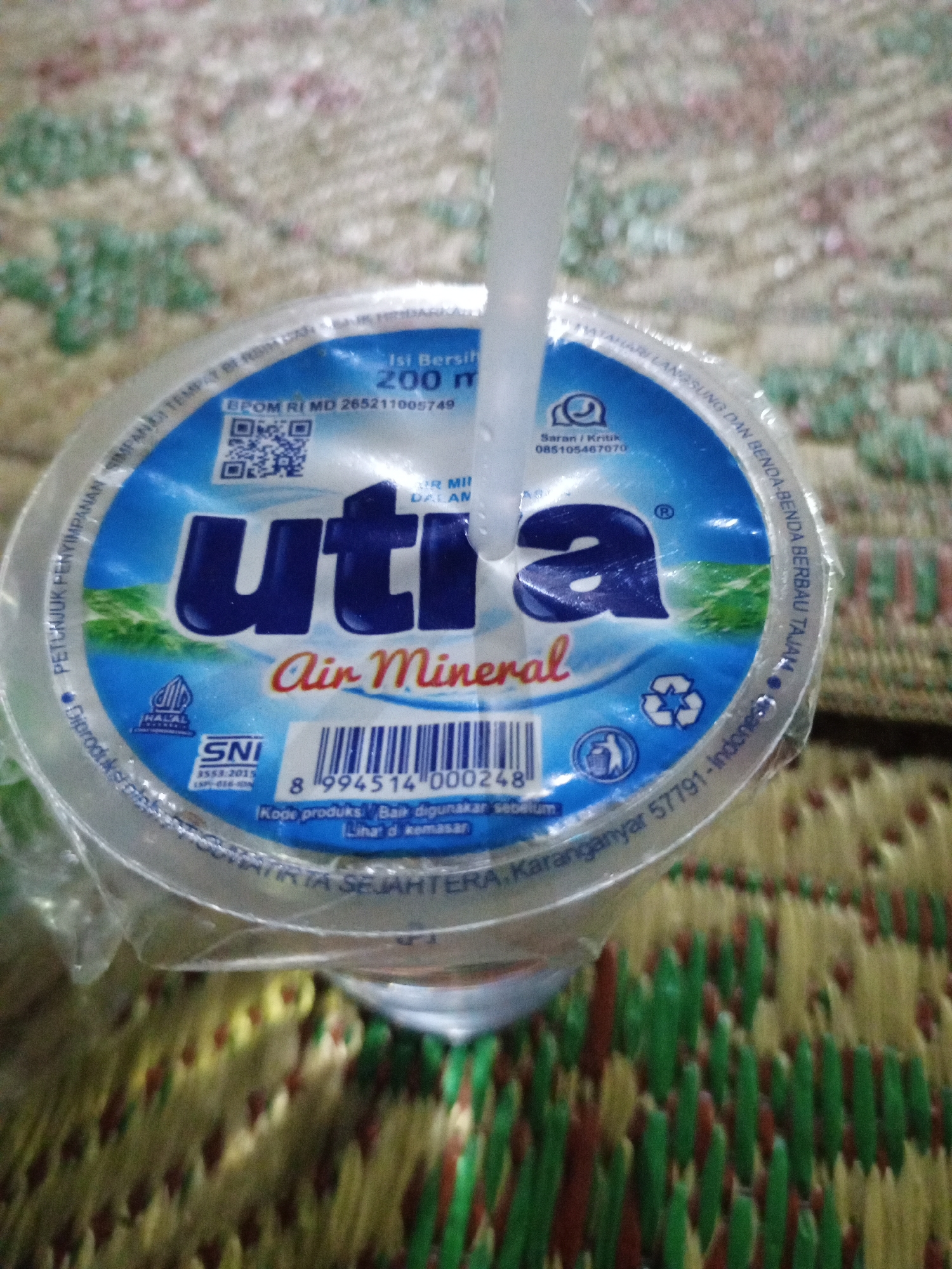 The image shows a close-up of a plastic cup of "Utra Air Mineral," a brand of bottled water, with a straw inserted. The cup has a blue and white label with text in Indonesian. The label indicates the content is 200 ml of mineral water and includes a barcode, Halal certification, and SNI (Standar Nasional Indonesia) certification. The surrounding area is a textured surface, possibly a mat or fabric, with a blurred background of green, red, and yellow. The text "PT. Tirta Sejahtera Karanganyar 57791 - Indonesia" suggests the product is manufactured by PT. Tirta Sejahtera in Karanganyar, Indonesia. Karanganyar is a regency in Central Java, Indonesia. The specific location within the city of Lemahbang is not identifiable from the image.