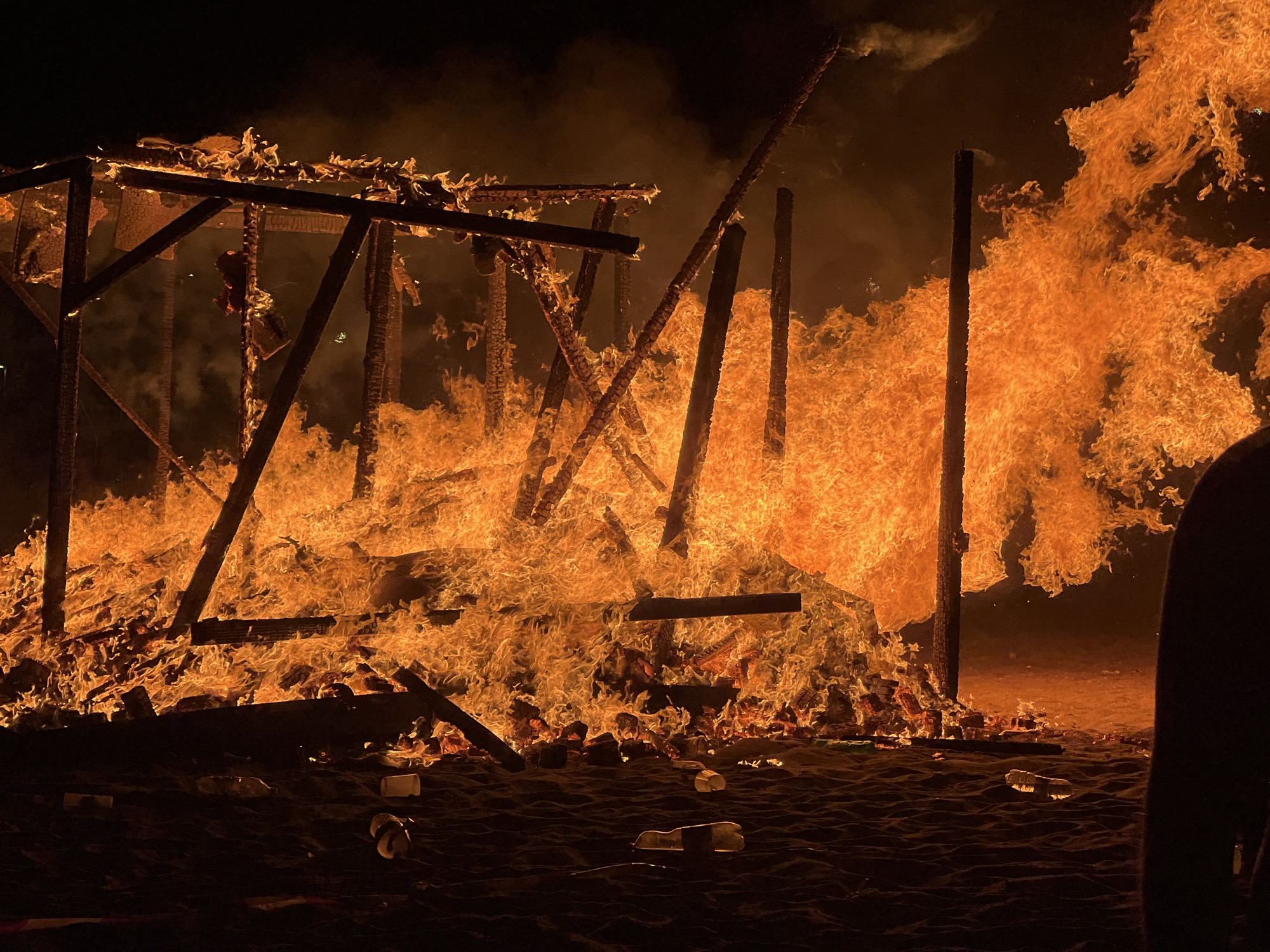 An open-air structure, possibly a temporary construction, is engulfed in intense flames. The fire is consuming the wooden beams and supports, creating a dynamic and chaotic scene with a significant amount of smoke and heat. The flames are primarily orange and yellow, with some white areas indicating extreme heat. The ground appears to be sandy, with several plastic bottles and cups scattered across it, suggesting a public event or gathering. The background is dark, indicating nighttime. The overall impression is one of destruction and intense heat. The image appears to have been taken in the city of La Línea de la Concepción, Spain.