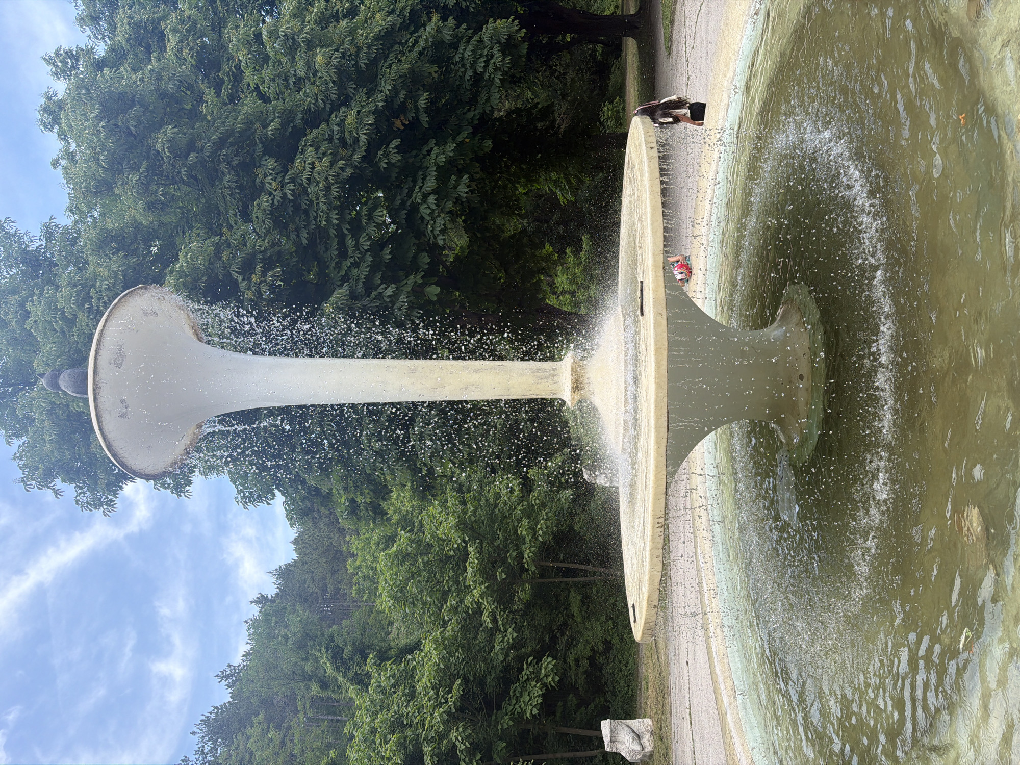 This image captures a multi-tiered concrete fountain in a park setting within the city of Shumen, Bulgaria. The fountain's central element is a tall, slender column supporting a wide, shallow upper basin. Perched on the top edge of this basin is a small, dark, stylized statue, possibly resembling an owl or a similar creature. Water cascades downwards from the upper basin in a continuous sheet of droplets, splashing into a broader, circular middle basin. From this middle basin, water overflows into a large, circular pool at ground level. The water in the lowest pool is murky, exhibiting a greenish-brown tint, suggestive of algae or sediment.
A small, dark stick or piece of debris rests on the edge of the middle basin. Floating debris is also visible in the ground-level pool. The fountain is surrounded by dense green foliage. To the left, a forested hillside dominates the background, composed of various deciduous and coniferous trees. To the right, large, mature trees with full green canopies frame the scene. Overhead, the sky is partially cloudy with discernible white, wispy clouds. A paved or dirt path encircles the fountain.
On the ground to the left of the fountain, near the treeline, an abstract, light-colored stone sculpture or monument is partially visible. On the right side of the fountain, a young woman with long dark hair, wearing a light-colored top and dark skirt or shorts, stands facing the fountain. She holds a pink or purple object, possibly a bag strap or racket handle, over her right shoulder. Near the fountain's edge on the right, a small red and white object, resembling a child's toy or deflated bag, lies on the ground.