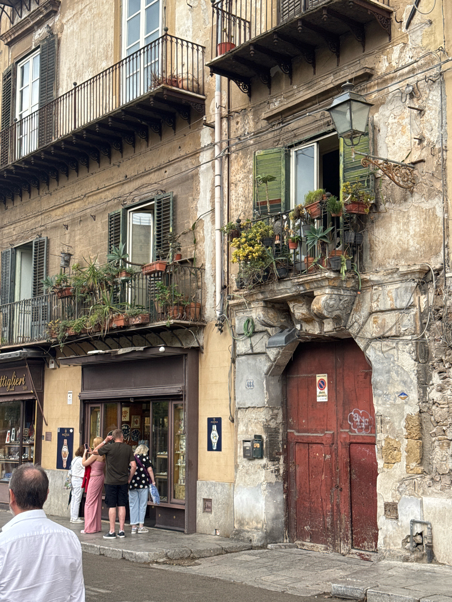 The image depicts a street scene in Palermo, Italy, focusing on the architecture of old buildings and a group of people interacting in front of a shop. The buildings feature weathered facades with balconies adorned with plants. On the right side, a large, weathered red door is prominent, set within an archway of crumbling stone. A street lamp is visible on the upper right of the building.
In front of a shop with a display window, four individuals are present. One person, facing away from the camera and wearing a white shirt, appears to be observing the scene. Another group of three people stands in front of the shop: a woman in a pink dress, a man in a dark shirt and shorts, and a woman in a polka dot top and jeans. The man is holding a phone up, possibly taking a photo. The shop itself appears to sell watches, as indicated by display items and signage. The street is paved with cobblestones and has a sidewalk. The overall impression is of an urban environment with historic buildings showing signs of age and wear, common in older European cities like Palermo.