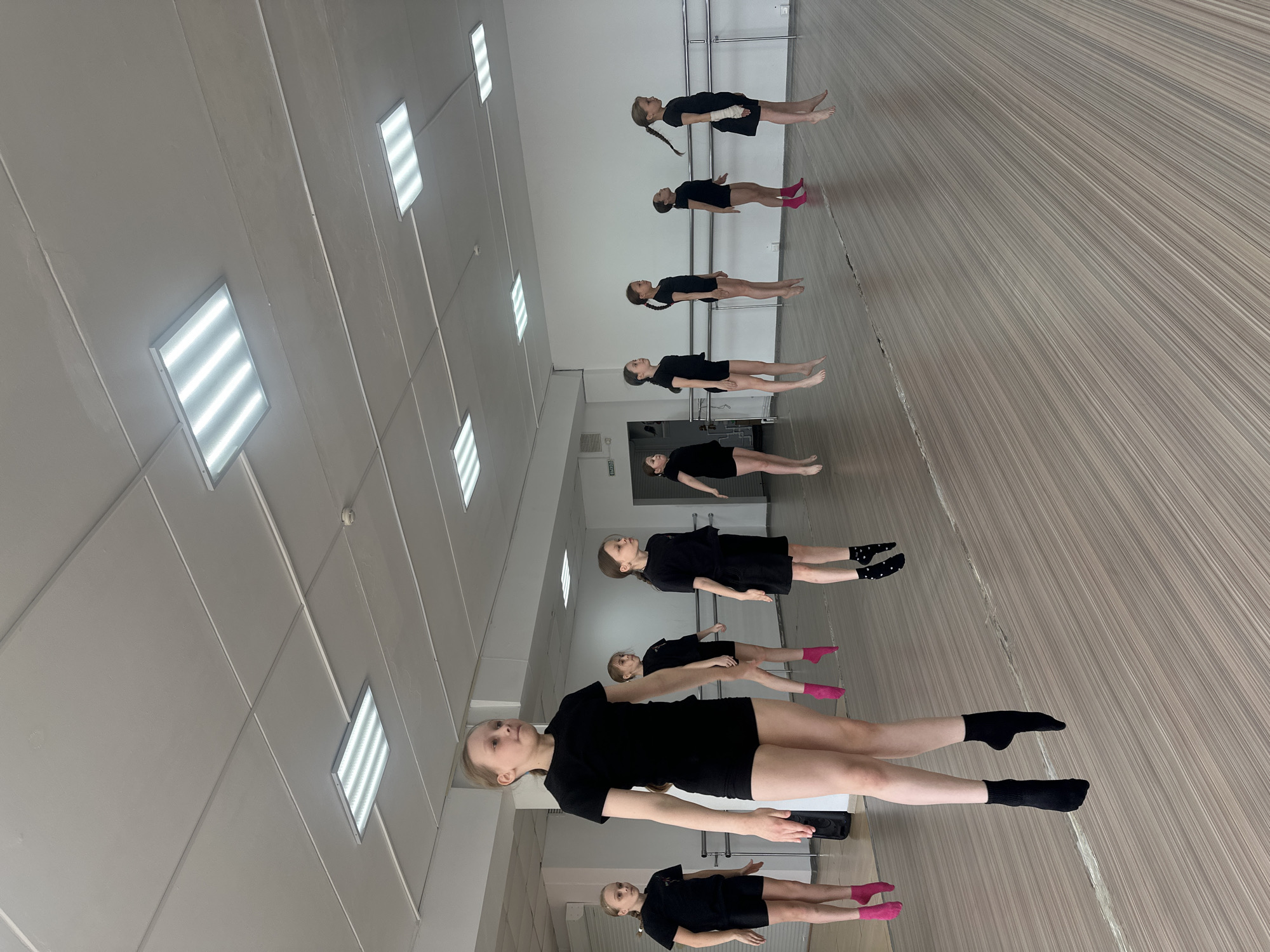A dance studio in Votkinsk, Russia, features a row of young girls lying on their backs on a light brown wood floor. They are all wearing black dance attire, some with pink or black socks. The room is illuminated by a series of rectangular fluorescent lights in the white ceiling, and a barre is visible along the wall in the background. The dancers are arranged in a line, with some looking up at the ceiling.