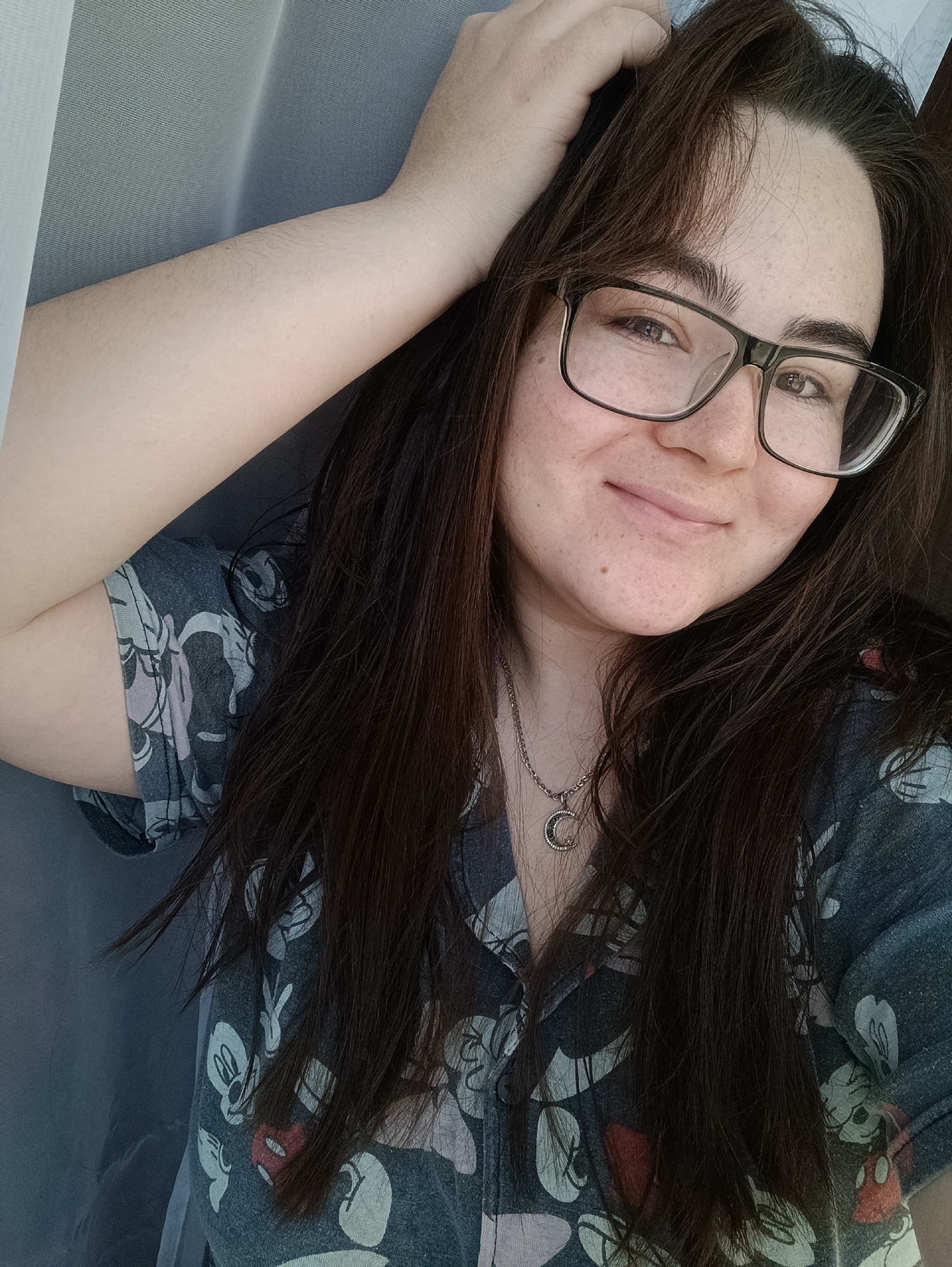 A woman with long, dark brown hair, wearing glasses and a gray shirt patterned with cartoon characters, is smiling at the camera. She has her right hand on her head and a silver necklace with a crescent moon pendant around her neck. She appears to be indoors, possibly near a window covered with a sheer curtain. There's no information to determine if this image was taken in Noviy, Russia.