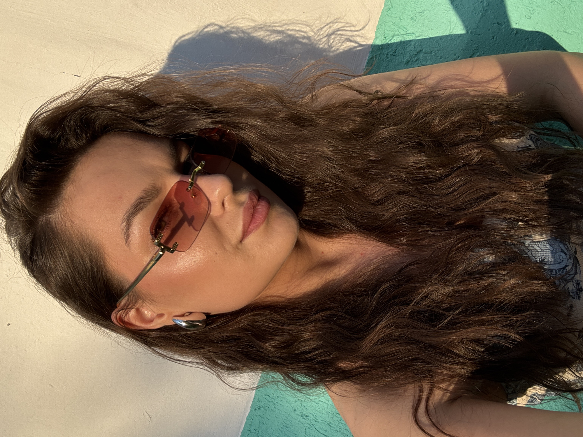A woman wearing large, rectangular, rimless sunglasses with pink lenses is leaning against a wall. She has long, wavy, brown hair that covers part of her face and shoulders. She is wearing a silver hoop earring in her left ear. The wall behind her is painted in two horizontal sections: the top section is white and the bottom section is a textured turquoise. A shadow of what appears to be a leafy branch is cast on the wall behind her. The lighting suggests it is late afternoon or early evening. No specific location in the city of Chaikovsky, Russia can be determined from the image.