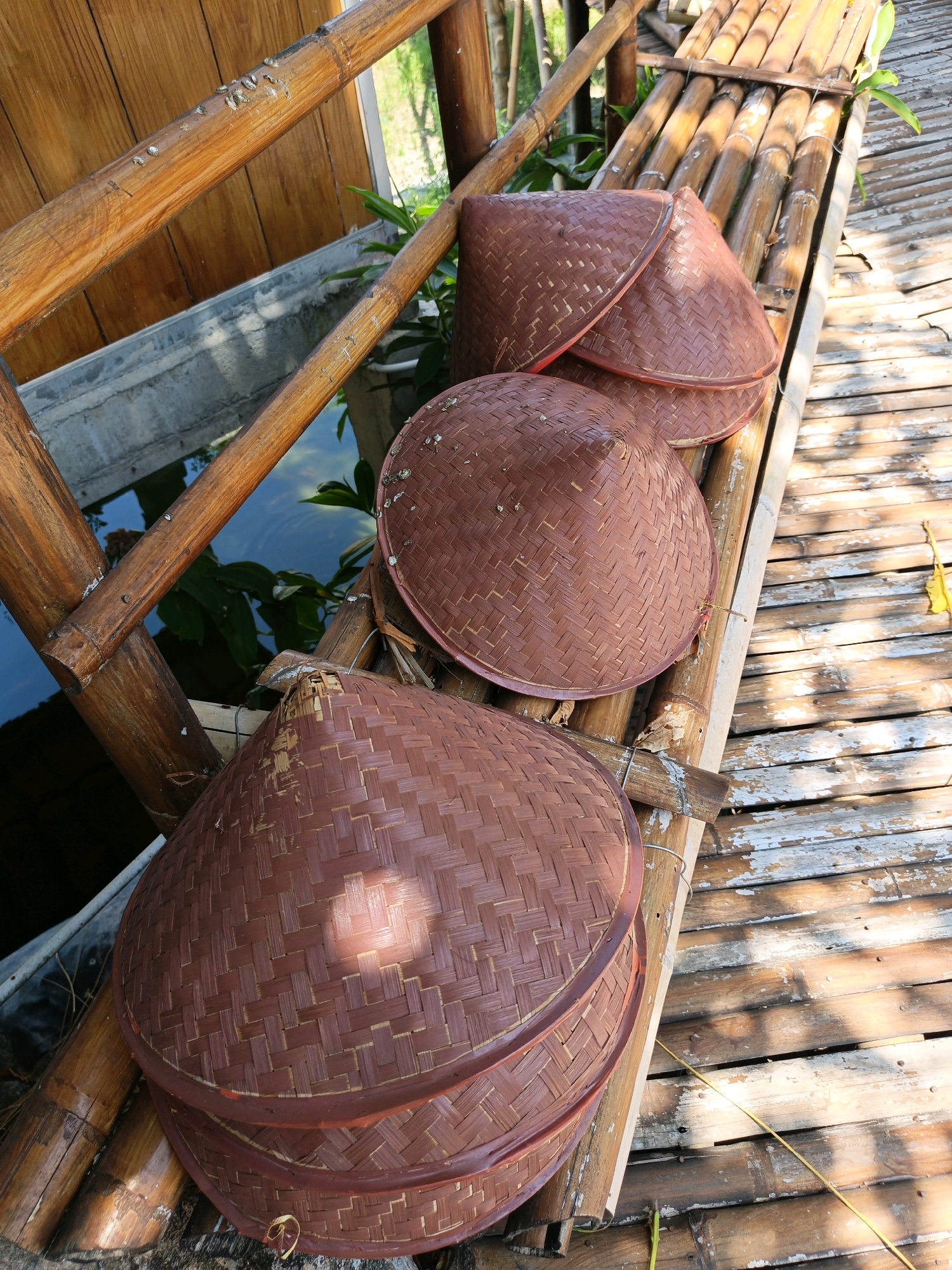 The image captures a collection of conical, woven bamboo hats (known as *caping* in Indonesia) resting on a bamboo structure. These hats are stacked and arranged on a slatted bamboo platform that appears to be part of an outdoor seating area or walkway. The bamboo elements are weathered, with some showing signs of age and wear. To the left of the hats, a concrete trough containing water and lush green foliage is visible, suggesting a garden or water feature. The background features a wooden wall and the dappled sunlight creates shadows on the bamboo flooring. The scene is likely located in Jonggrangan, Indonesia, given the cultural context of the *caping* and bamboo construction. There are no people or animals visible in the image.