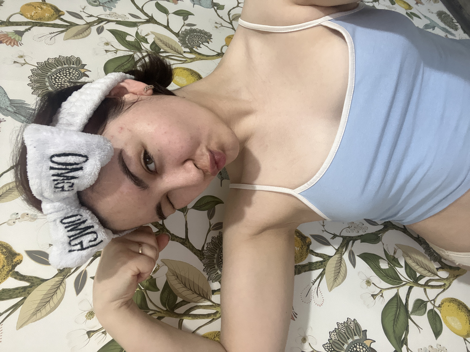 A woman with dark hair pulled back into a bun is lying on a floral-patterned surface. She is wearing a light blue tank top with white straps and a white terrycloth headband with the word "OMG" printed on it. She is making a puckered facial expression, and her right arm is bent with her fist near her face. There is a ring on her left ring finger. The background features a print with lemons, leaves, and flowers. The location in the city of Neftekamsk, Russia is unknown.