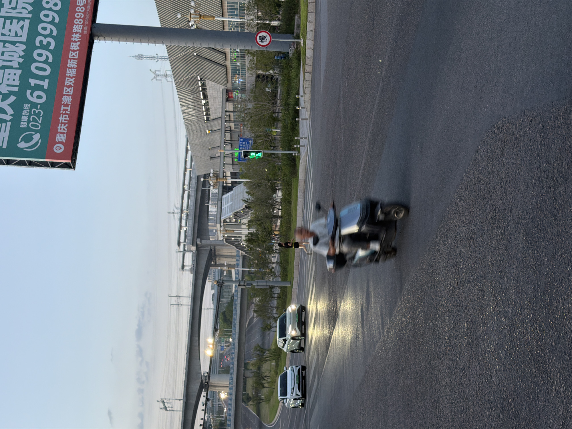 The image depicts an urban scene in Jiangjin, China, at dusk. A road in the foreground is wet from recent rain, reflecting the artificial lights and the sky. Two white cars are driving on the road, with headlights illuminated. A person is operating a three-wheeled scooter, moving from right to left across the frame, their movement blurred due to motion. In the background, a multi-level elevated roadway passes over the scene, with traffic lights visible. A modern building with a distinctive angular roofline is situated behind the roadway. A large billboard advertising "Chongqing Fucheng Hospital" is prominently displayed in the upper right quadrant of the image, including a phone number and an address in Jiangjin District. A traffic sign with the Chinese character "慢" (màn, meaning "slow") is visible on a pole to the right of the building. The sky is a pale blue, indicating the transition from day to night.