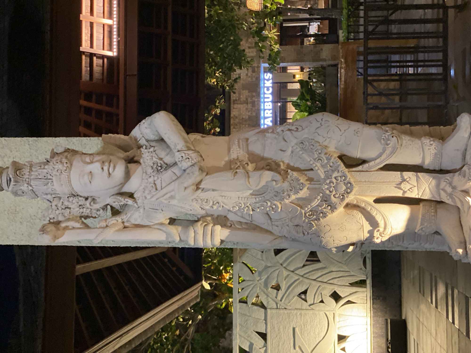 A nighttime exterior shot shows a tall, intricately carved stone statue of a figure wearing ornate headdress and clothing, positioned in front of a building. The statue is illuminated by a warm light from the left. Behind the statue, a building with a distinctive neon "STARBUCKS" sign is visible, along with trees and foliage. A thatched roof structure is seen above and to the left of the statue. The foreground features a tiled walkway and a decorative stone wall with leaf motifs. The overall setting appears to be an outdoor area of a commercial establishment in Kuta Selatan, Indonesia.