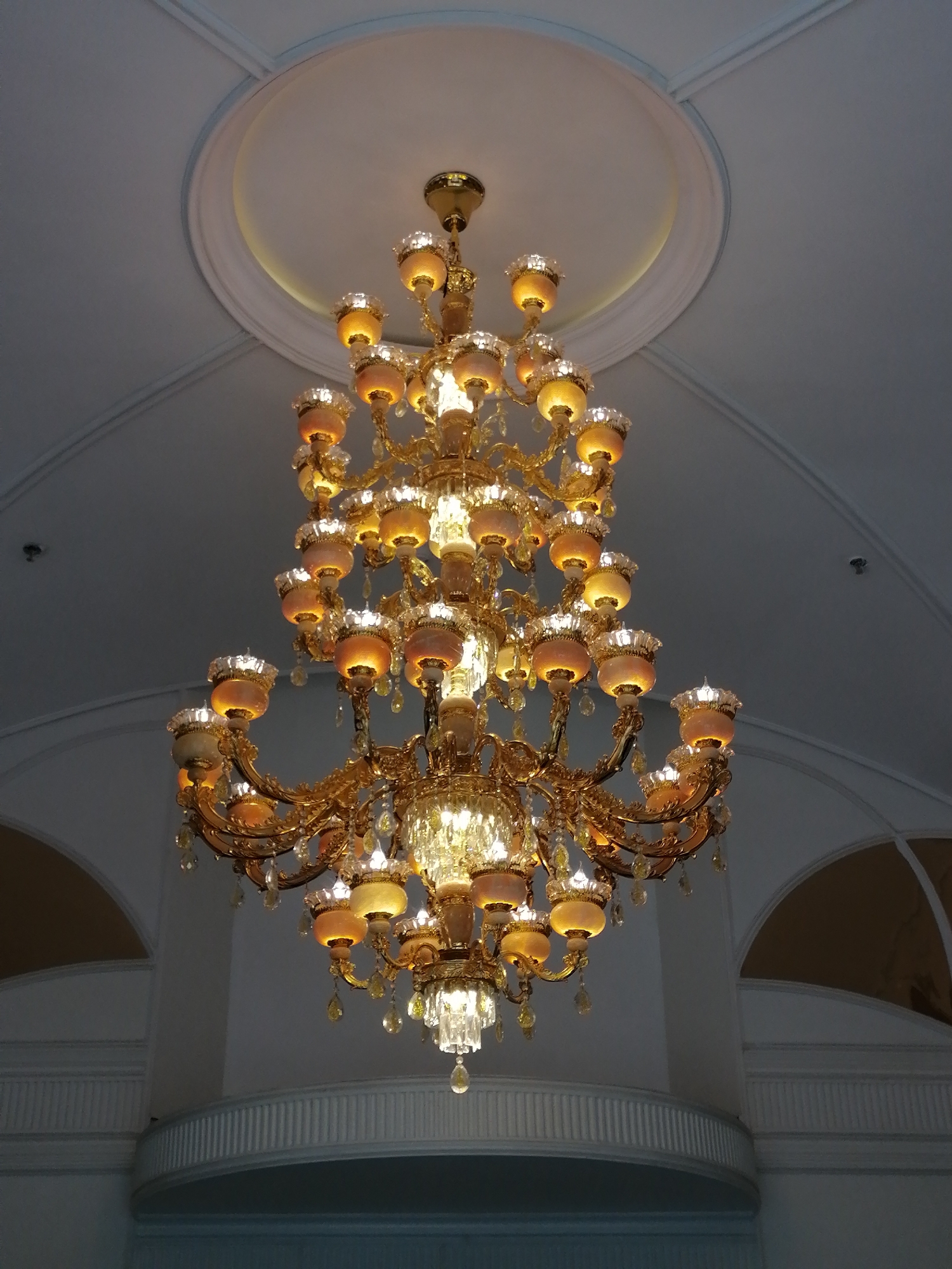 The image shows a large, ornate chandelier hanging from a ceiling. The chandelier is gold-colored with multiple tiers of lights. The lights appear to be enclosed in translucent shades, possibly glass or frosted plastic. The ceiling is white with a recessed circular design around the chandelier's base. The location appears to be indoors, possibly a grand hall or ballroom. There are architectural details in the background, including arched windows or doorways. The scene is well-lit, with the chandelier providing the primary source of illumination. The location is in Sa Pa Ward, Vietnam. There are no people visible in the image.