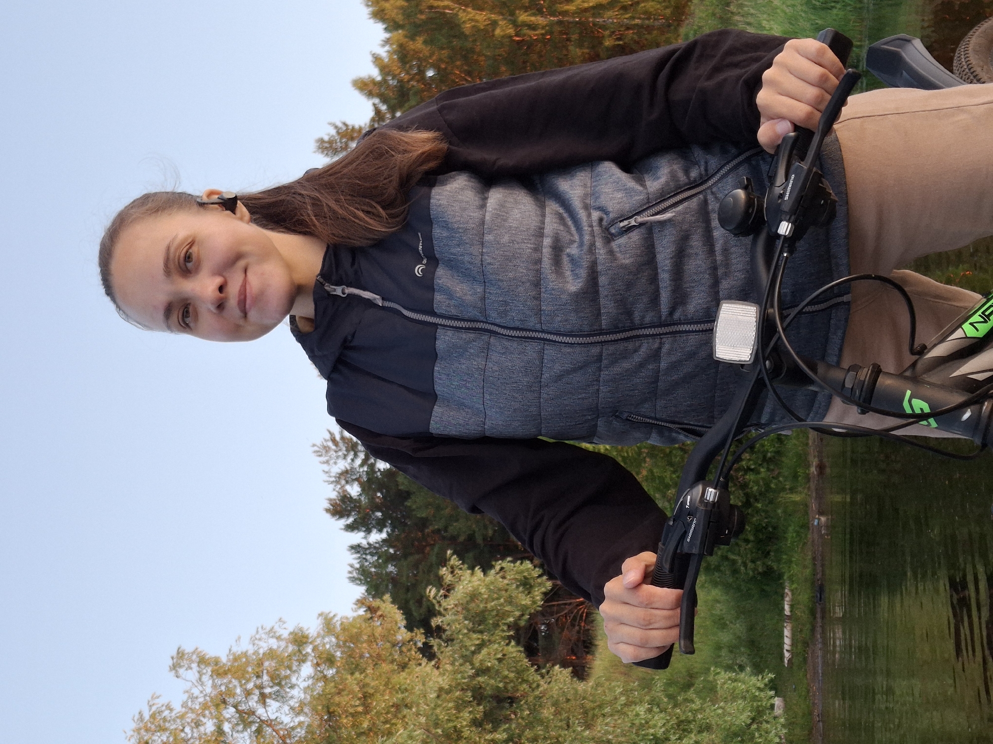 A medium shot captures a woman with dark hair pulled back in a ponytail, wearing a grey and black hooded vest and tan pants, standing with a bicycle. She has a slight smile and is looking towards the camera, with a black wireless earbud in her right ear. Her hands are on the handlebars of the bicycle. The background features a body of water with reflections, and trees with foliage in various shades of green and yellow. The sky is clear and light blue, suggesting it is daytime. The scene appears to be outdoors, possibly near a lake or river, with a backdrop of a natural environment. The city of Chaikovsky, Russia, is known for its scenic locations along the Kama River, and this image aligns with such a setting, suggesting it could be captured there.