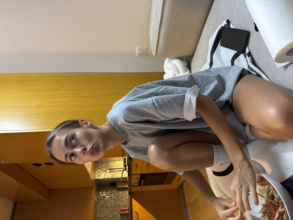 A woman wearing a grey oversized shirt and white socks squats indoors, appearing playful. She is positioned near a sofa with a cell phone and paper towels on it. In front of her, she holds a plate of food, possibly crab or lobster. The background includes a kitchen area, with cabinets and appliances visible. The overall scene appears casual and domestic.