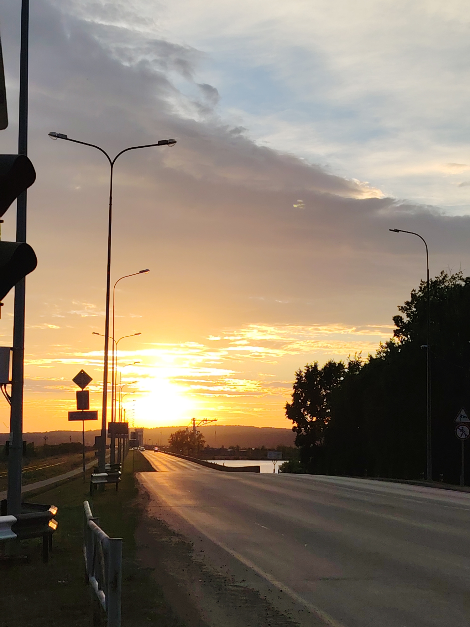 The image shows a road leading towards a sunset in the city of Chaikovsky, Russia. The sky is filled with colorful clouds, with the sun appearing as a bright orange ball on the horizon. The road is wet and reflects the colors of the sunset. There are streetlights along the road, and trees line the right side of the image. In the distance, there appears to be a body of water. The scene is peaceful and serene, with no people or animals visible. The image appears to have been taken from a slightly elevated position, possibly from a bridge or overpass, looking down the road.