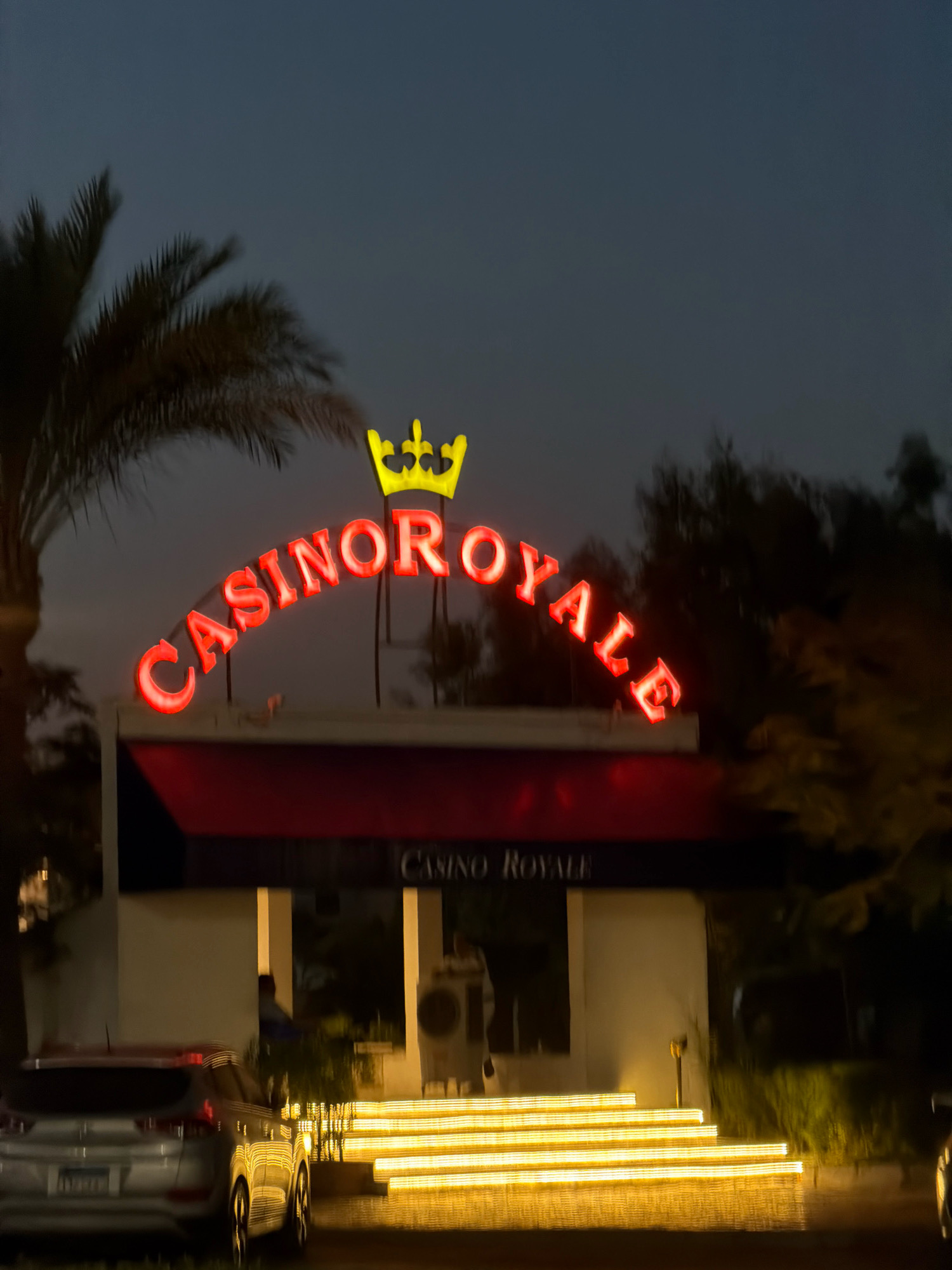 A neon sign with red letters that spell "CASINO ROYALE" is illuminated against a dark sky. A yellow crown sits above the sign. In front of the sign is a building with a red awning. A set of illuminated stairs leads up to the building. A silver SUV is parked in front of the stairs. The scene is likely at night, given the dark sky and illuminated signage. The presence of palm trees suggests a warm climate, and the architectural style and signage may indicate a tourist or entertainment district in Sharm El Sheikh, Egypt. There are no discernible people clearly visible or identifiable in the scene, although blurry figures may be present in the background within the building.