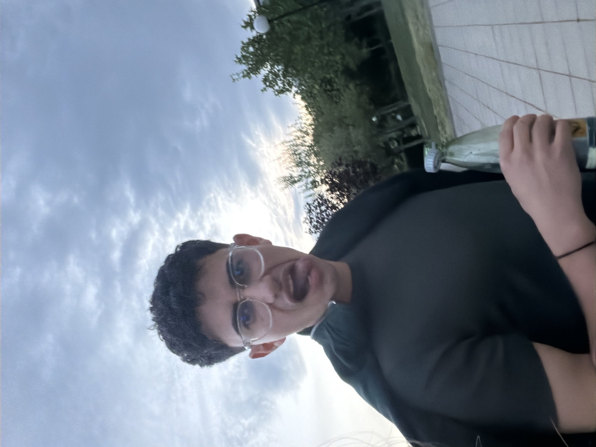 A young man with dark curly hair and glasses is posing for a selfie. He is wearing a dark-colored shirt or jacket. He is making a silly face, sticking out his tongue slightly. His right hand holds what appears to be a clear glass bottle. The background includes a cloudy sky with the sun breaking through, trees, and a structure that might be a railing or ledge. The location appears to be outdoors. While the image's setting is not sufficiently clear to ascertain definitively the city in the image, without more information it is not possible to assert that the image was taken in Blagoevgrad, Bulgaria.