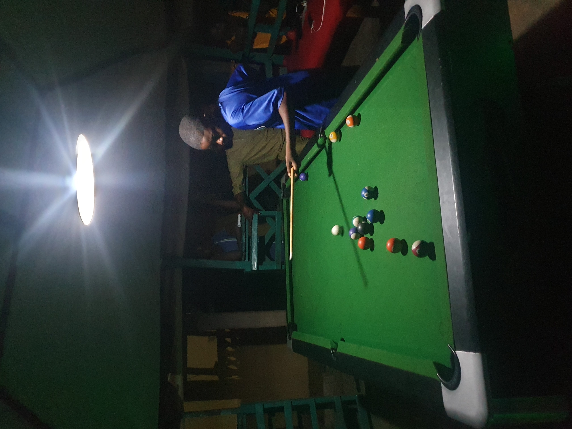 The image shows a person playing billiards in what appears to be an indoor setting. The person, dressed in a blue shirt and khaki pants, is holding a cue stick and aiming at the billiard balls on the green felt table. Several billiard balls are visible on the table, in various colors and numbers. The lighting in the scene is dim, with a bright light source, possibly a lamp, illuminating the area around the table. This suggests the game might be taking place at night or in a dimly lit establishment. The surroundings are somewhat dark, with glimpses of furniture or structures that indicate it could be a bar or a recreational area. The overall mood of the image seems to be focused and perhaps leisurely, given the activity. There is no visible text in the image.