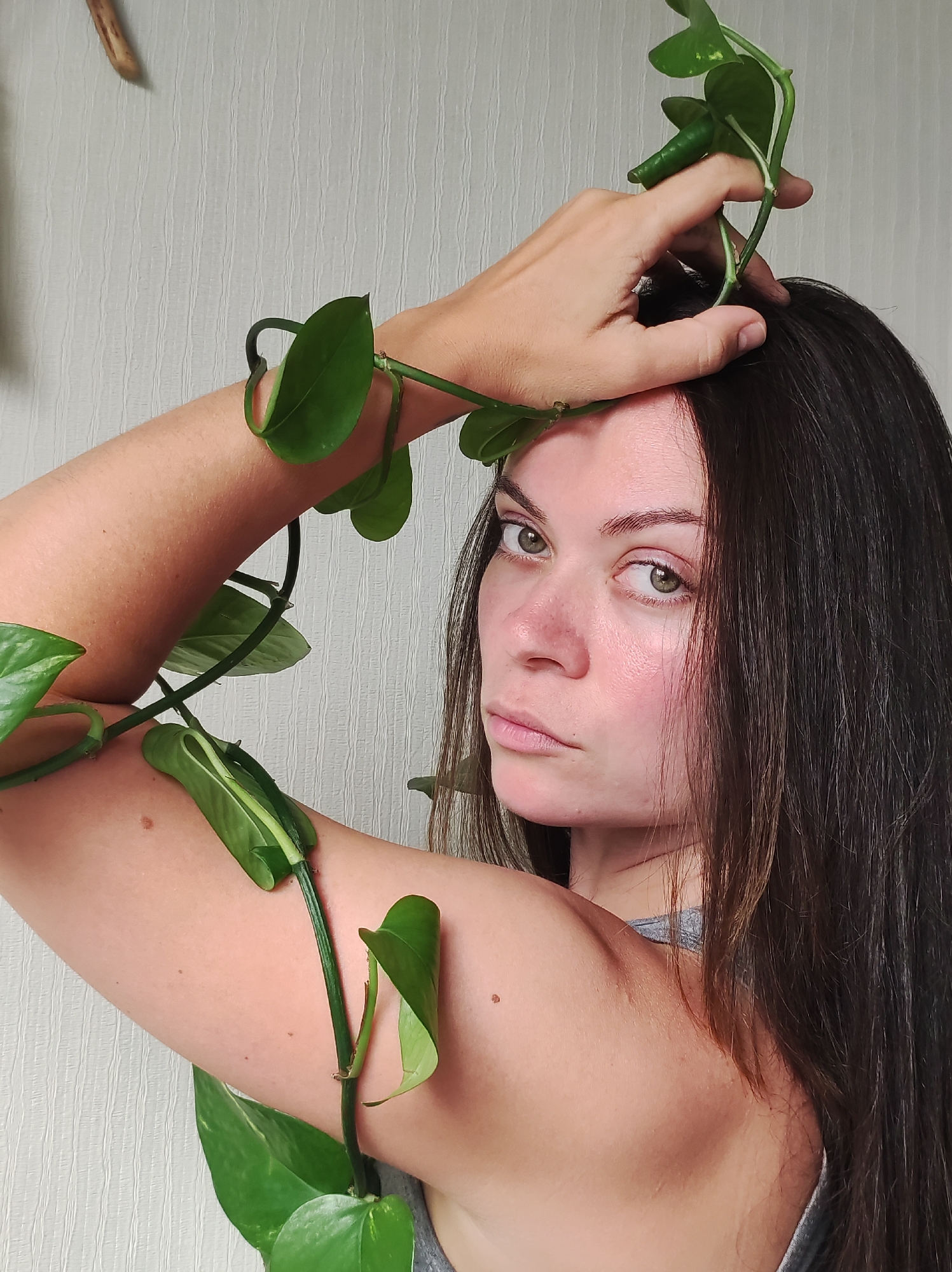 A young woman is depicted in a close-up, half-body portrait, looking directly at the viewer with a neutral and direct expression. She has long, dark brown hair, green eyes, and fair skin with some natural redness visible on her cheeks and nose. A vibrant green vining plant, with distinctive heart-shaped leaves, is intertwined around her left arm, creating a natural accessory. Her left hand is raised, resting on the top of her head, with more of the vine's leaves and stems draped over her hand and hairline. She appears to be wearing a simple grey top, with a strap visible near her right shoulder.
The background consists of a light-colored wall with a subtle, vertically textured pattern. In the upper left corner, a small, irregularly shaped light-brown wooden object, possibly a decorative twig, is visible against the wall. The lighting in the image is soft and even, suggesting an indoor setting, likely during the daytime. No text is visible in the image.
