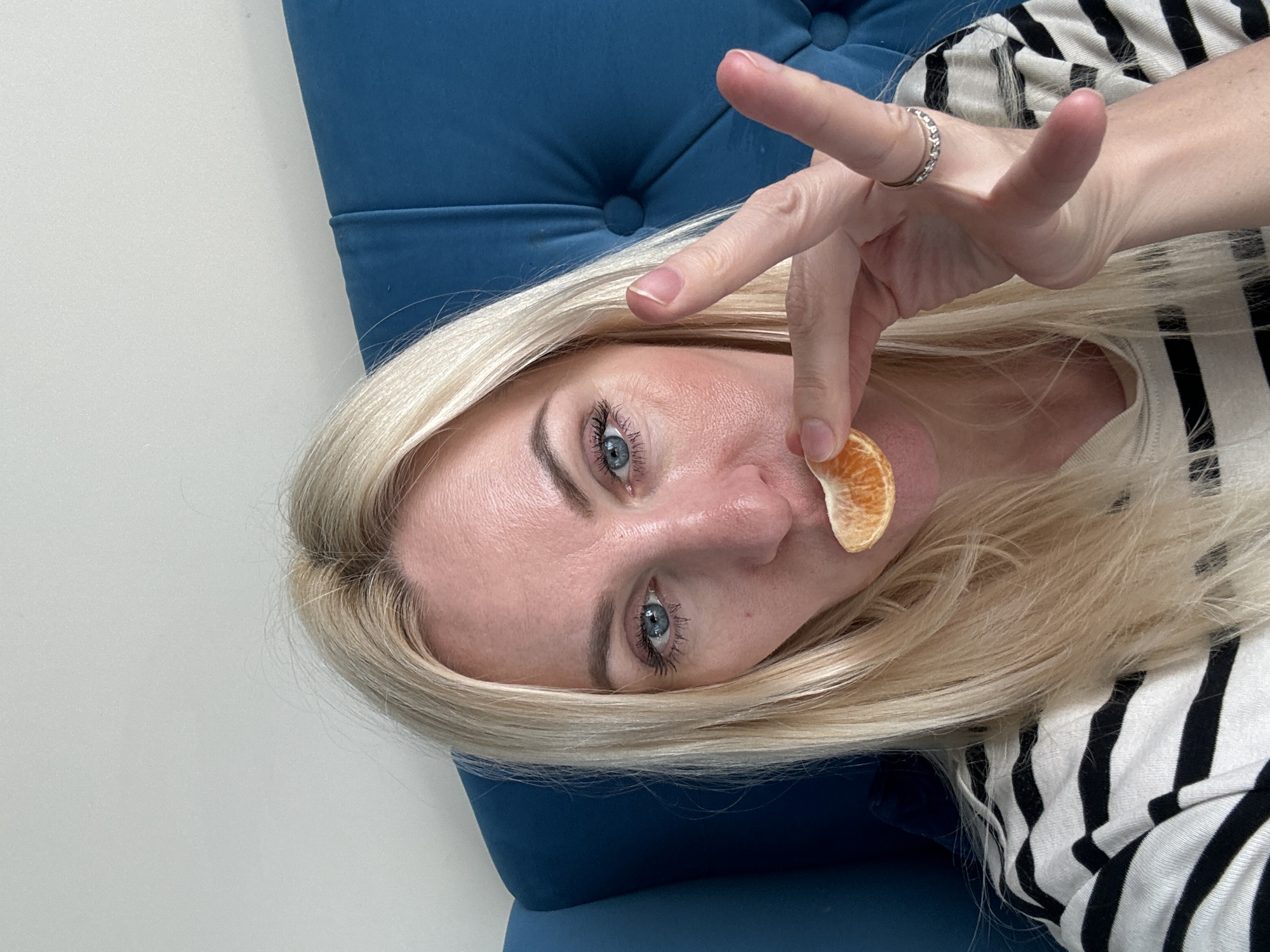 In a close-up, selfie-style shot, a fair-skinned woman with blonde hair is seated in what appears to be a tufted blue armchair against a plain white wall. She is holding a slice of tangerine up to her lips with her right hand, and her index and middle fingers are extended in a peace sign. A silver ring with small stones adorns her ring finger. She is wearing a black and white striped long-sleeved shirt. Her eyes are blue, and she has on mascara. The overall lighting suggests an indoor setting with natural light. The location is not specified in the image or context.