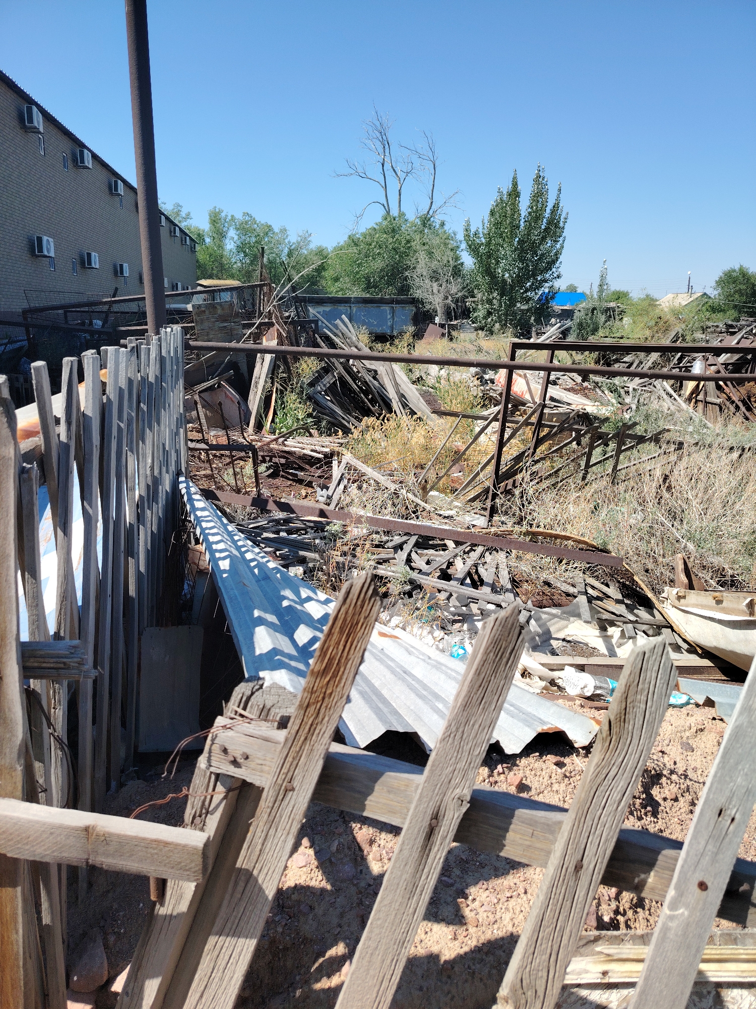 The image depicts a cluttered outdoor area, likely a junkyard or storage yard, in the vicinity of Тораңғылық, Kazakhstan. In the foreground, a weathered wooden fence constructed from vertical planks casts sharp shadows. Behind the fence, a chaotic jumble of scrap metal, including corrugated roofing sheets and various structural components, is piled high. Dry brush and weeds are interspersed throughout the debris. To the left, a portion of a large, multi-story brick building with numerous air conditioning units mounted on its facade is visible, suggesting an urban or semi-urban setting. The sky above is clear and blue. The scene is devoid of any people or animals. The overall impression is one of disarray and abandonment.