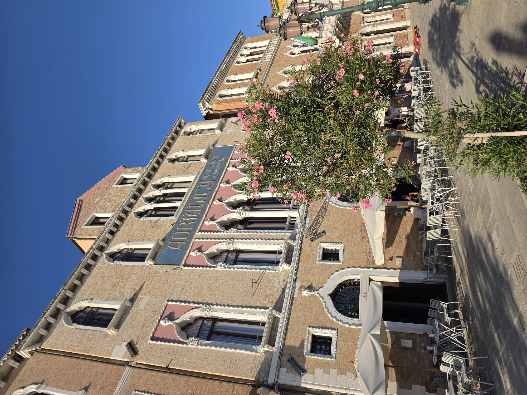 The photograph captures an exterior view of the Adriatica building in Venice, Italy, on a clear, sunny day. The building, constructed with light-colored bricks, features a classic Venetian architectural style with arched windows, ornate decorative elements, and a prominent blue sign with gold lettering that reads "ADRIATICA." Pink floral bushes are in bloom in front of the building, and several tables and chairs belonging to an outdoor cafe are set up on the cobblestone pavement. In the background, a streetlamp with multiple lights stands tall. People are visible seated at some of the tables. The scene conveys a serene and inviting atmosphere characteristic of Venice.