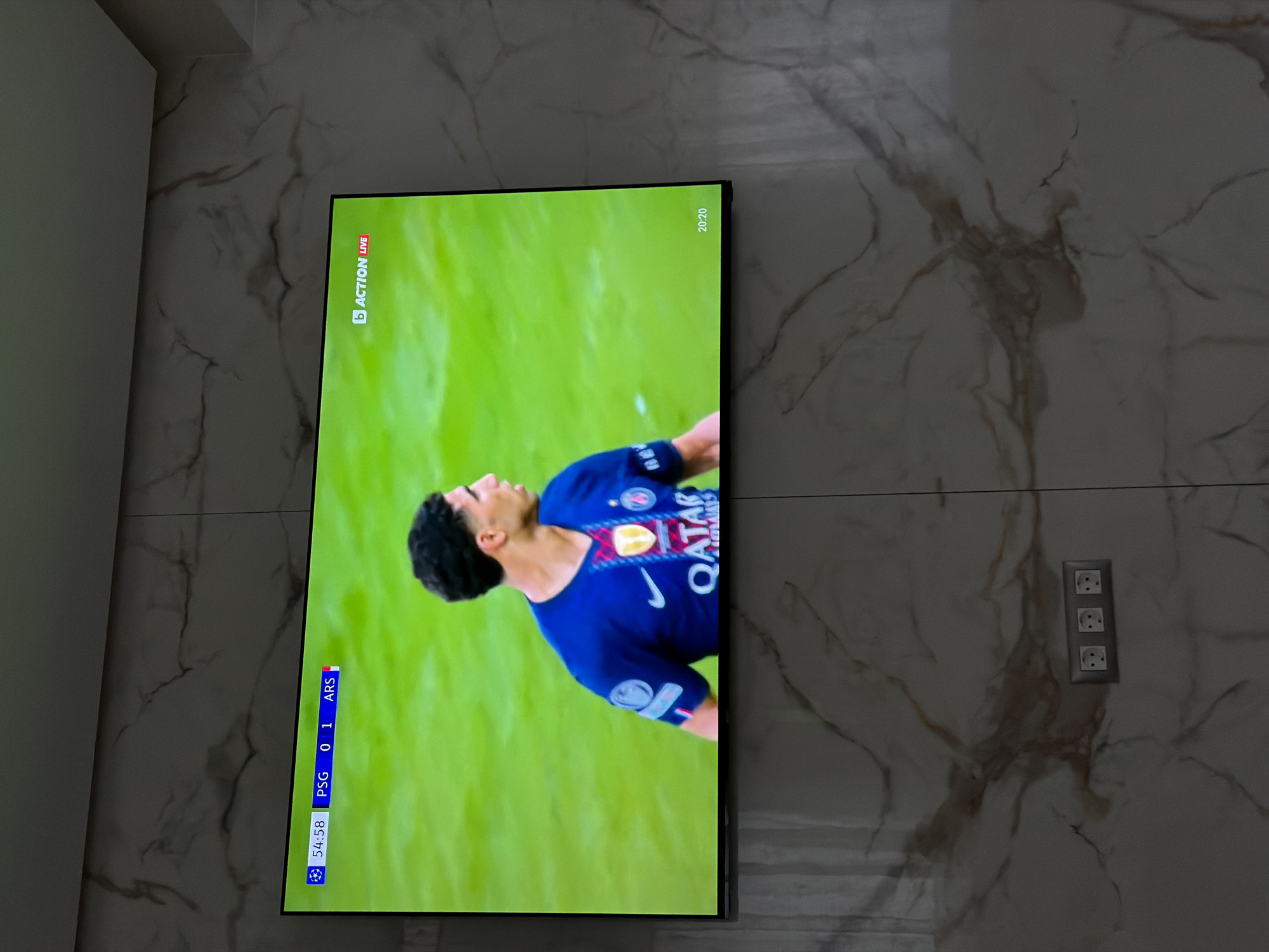 The image shows a television screen displaying a football match, PSG vs ARS, with the score 0-1. It is the 54th minute and 58 seconds of the game. A player in a blue Qatar branded shirt is visible on the screen. The TV is mounted on a marbled wall. A triple electrical socket is visible on the wall to the right of the TV. It is impossible to verify the location in Asenovgrad, Bulgaria, without further information.